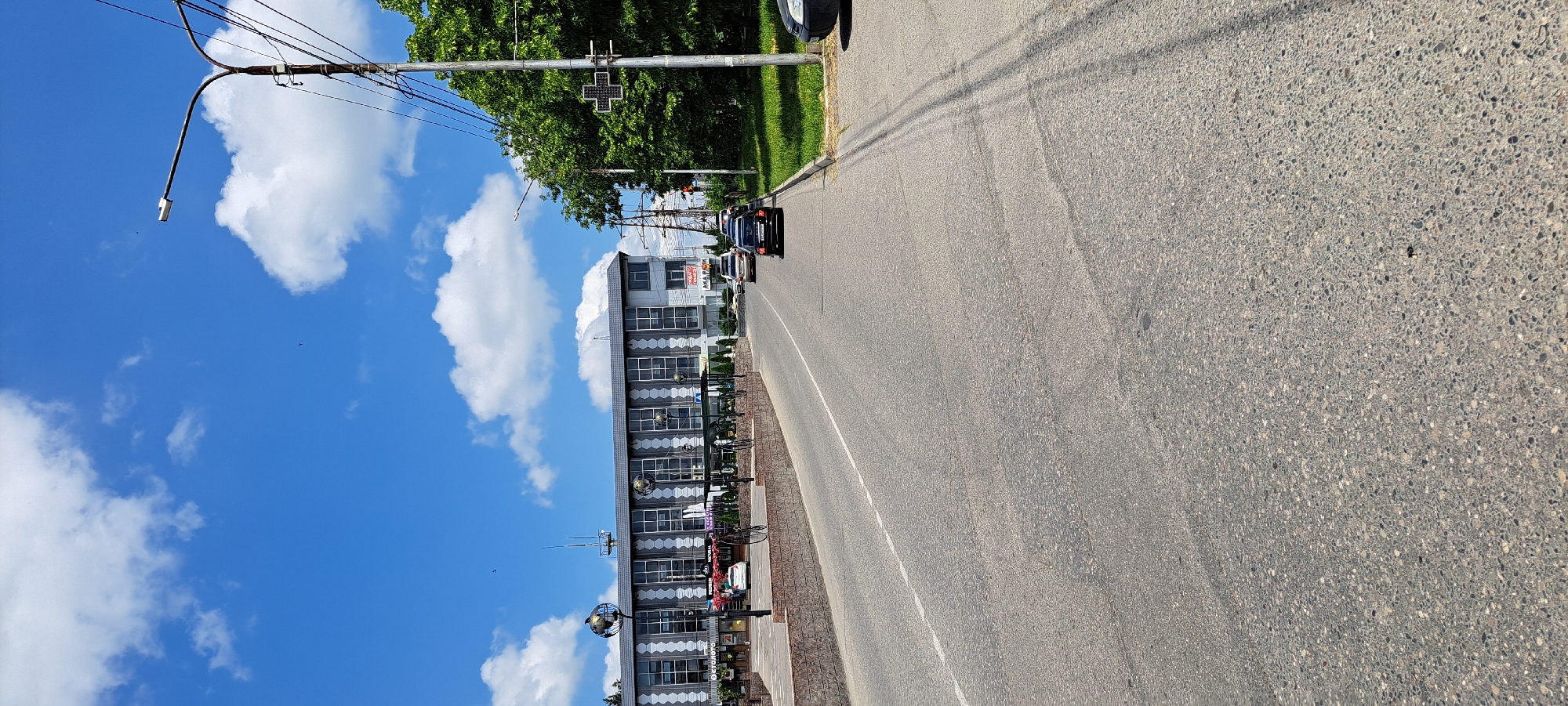 The image depicts a street scene in Yessentuki, Russia, under a bright blue sky with scattered white clouds. A modern, multi-story building with a grey roof and a facade of alternating dark grey and white vertical stripes and large windows stands on the left side of the street. In front of the building, a low, brick-faced wall lines a paved area with decorative globe-topped lampposts and some greenery. A road with a white dashed line marking the lane separation occupies the foreground and stretches into the distance. Several cars are visible on the road, including a black car in the middle lane and a silver car further ahead. On the right side of the road, there is a grassy area and a tall lamppost with electrical wires attached. A traffic signal displaying a red cross shape is also mounted on a pole on the right. The overall impression is a typical urban street on a clear day.