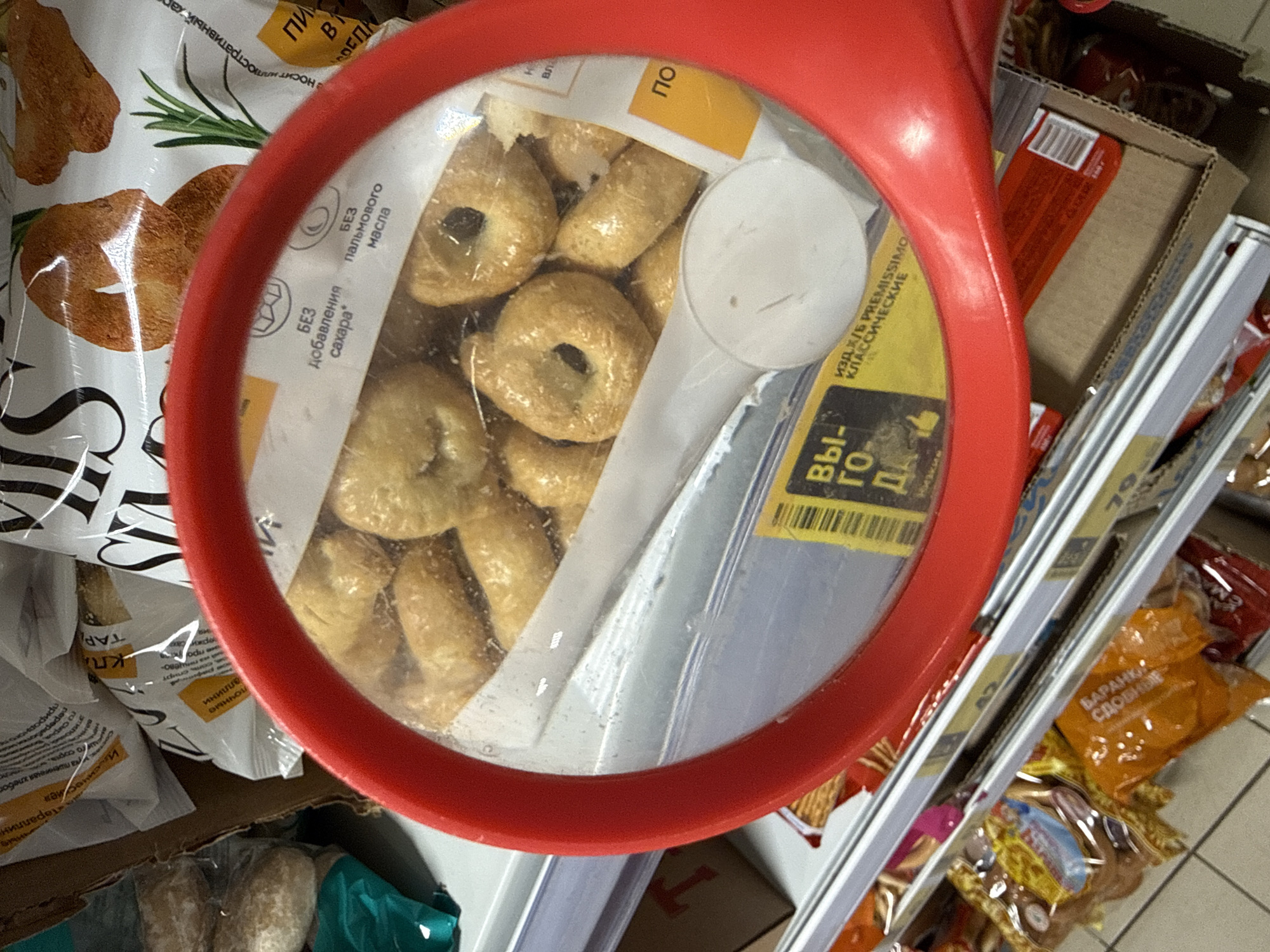 The image captures a shelf in a store, likely a supermarket, in Dmitrov, Russia. The focal point is a package of small, ring-shaped crackers or biscuits, possibly "Baranki Sdobnye" as indicated on a nearby product. The crackers are in a clear plastic container, partially obscured by a white plastic spoon or scoop. The package is seen through a red magnifying glass. The crackers are positioned on a shelf alongside other packaged goods, including other cracker or biscuit varieties. Text on some packages is in Russian. The environment appears to be a typical retail space, with shelved products, suggestive of a shopping environment.