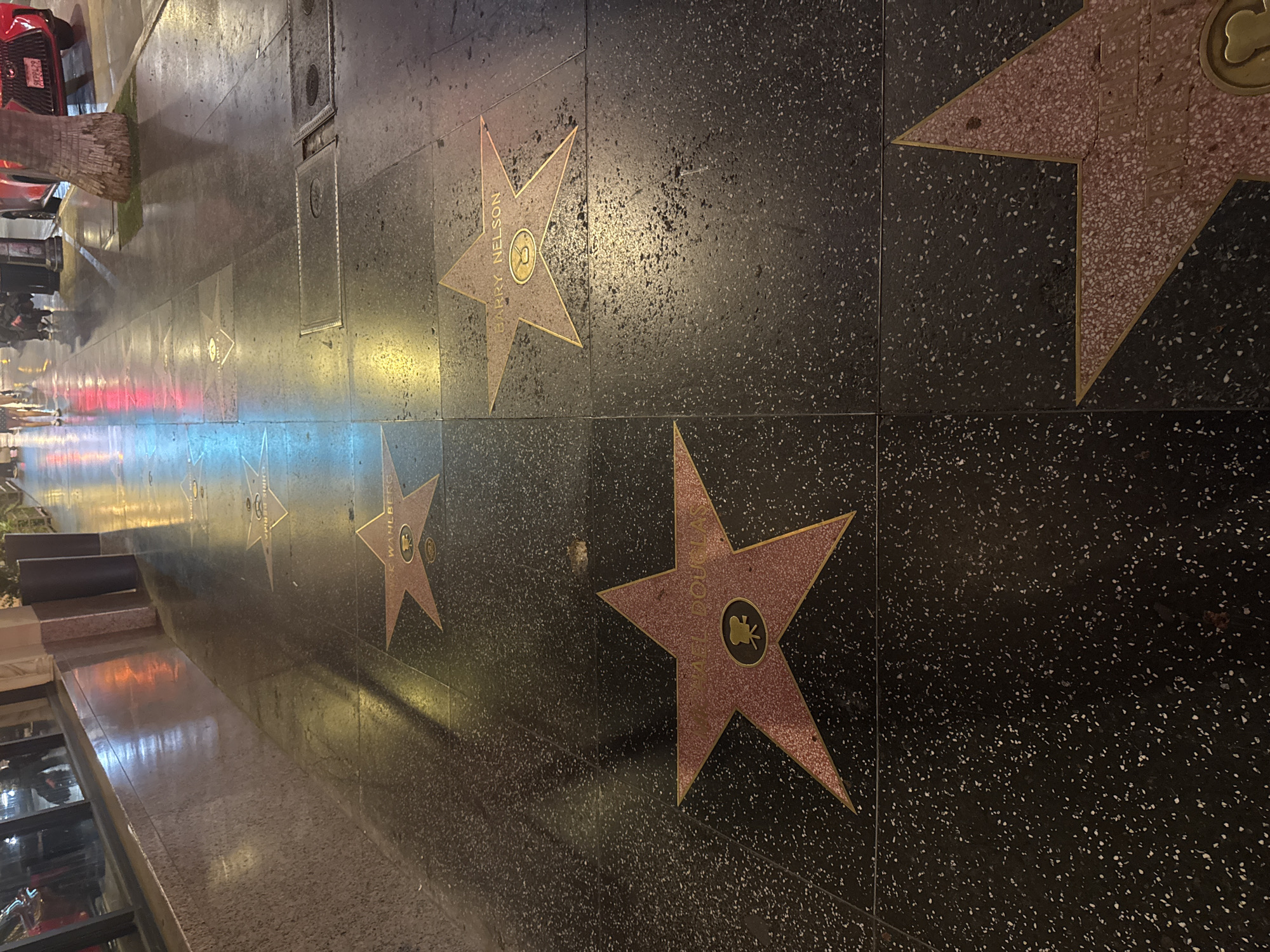 The image displays a section of the Hollywood Walk of Fame in Los Angeles. Paved with black terrazzo inlaid with gold stars, the walkway features the names of several honorees. Visible stars include "MICHAEL DOUGLAS" with a film camera emblem and "BARRY NELSON" with a vintage microphone emblem. The wet surface of the walkway reflects the ambient lights from nearby buildings, creating a shimmering effect. In the background, the reflections of neon signs and streetlights are visible on the wet pavement. People are walking along the sidewalk in the distance. To the right, a tree trunk and a red car are partially visible. The lighting suggests it is evening or night.