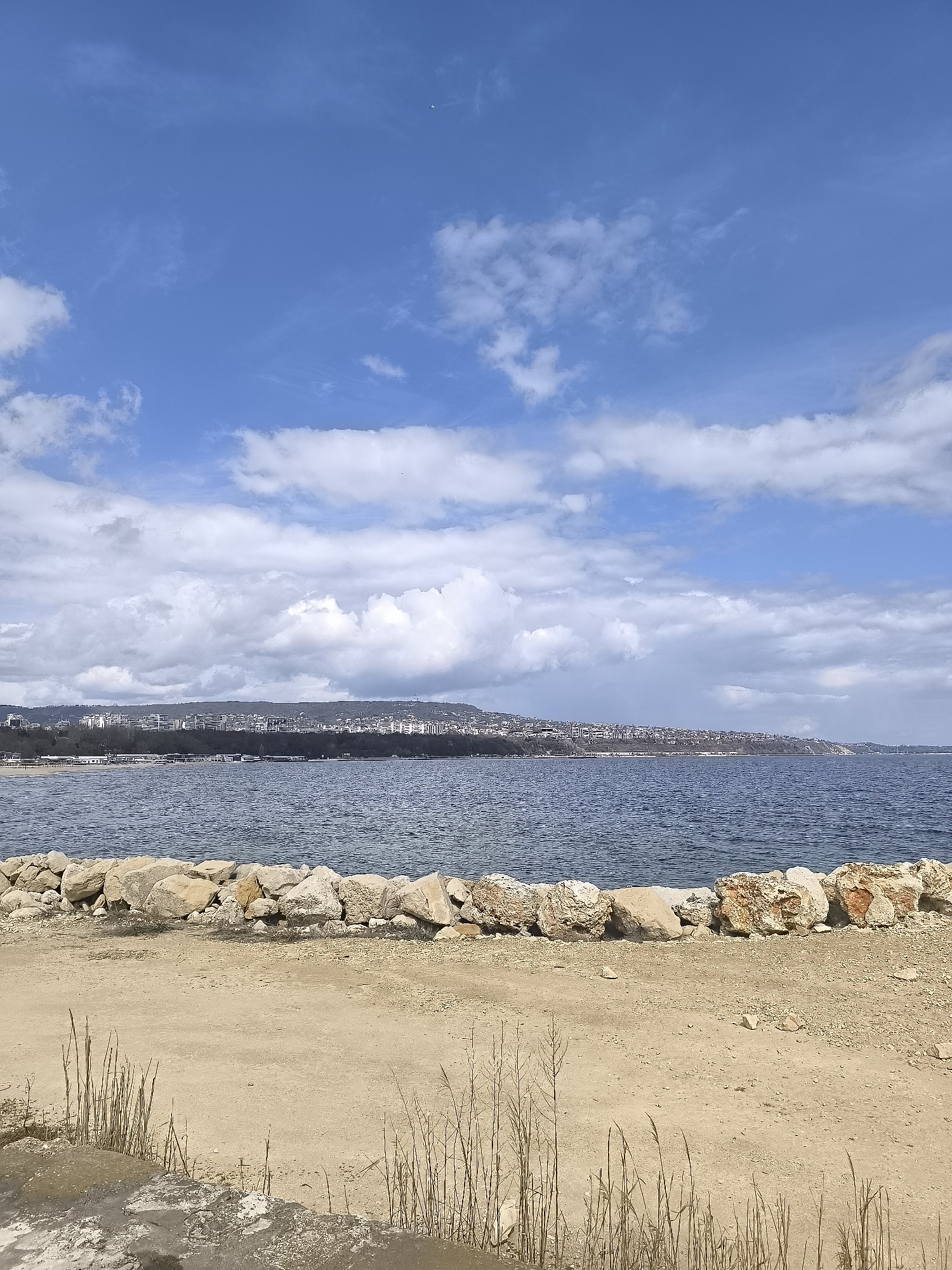The image shows a landscape scene in Varna, Bulgaria. In the foreground, there is a beach with a sand and gravel surface and some sparse vegetation. A barrier of rocks runs along the edge of the beach, separating it from the water. The water appears calm and stretches out toward the horizon. In the background, there is a coastline with buildings and trees, suggesting an urban area. The sky is blue with white clouds, indicating a partly cloudy day. There is no sign of human activity, such as people or moving vehicles.