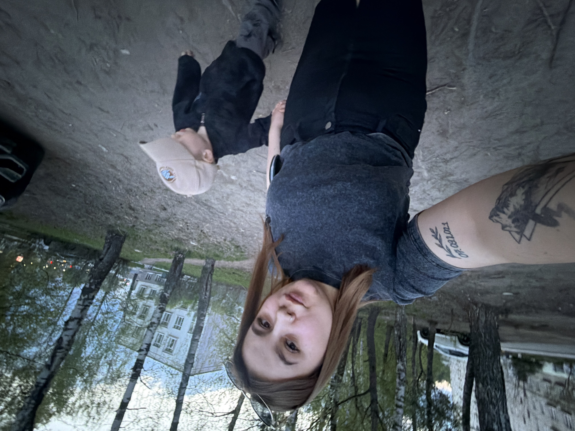 A woman takes a selfie with a child, likely a son, outside in Chaikovsky, Russia. They stand on a gravel or dirt surface. Behind them are birch trees and a multi-story residential building. The woman wears a grey t-shirt and black pants. She has a tattoo on her arm. The child wears a dark jacket and a tan baseball cap. A car is partially visible in the background. The image is rotated counter-clockwise.