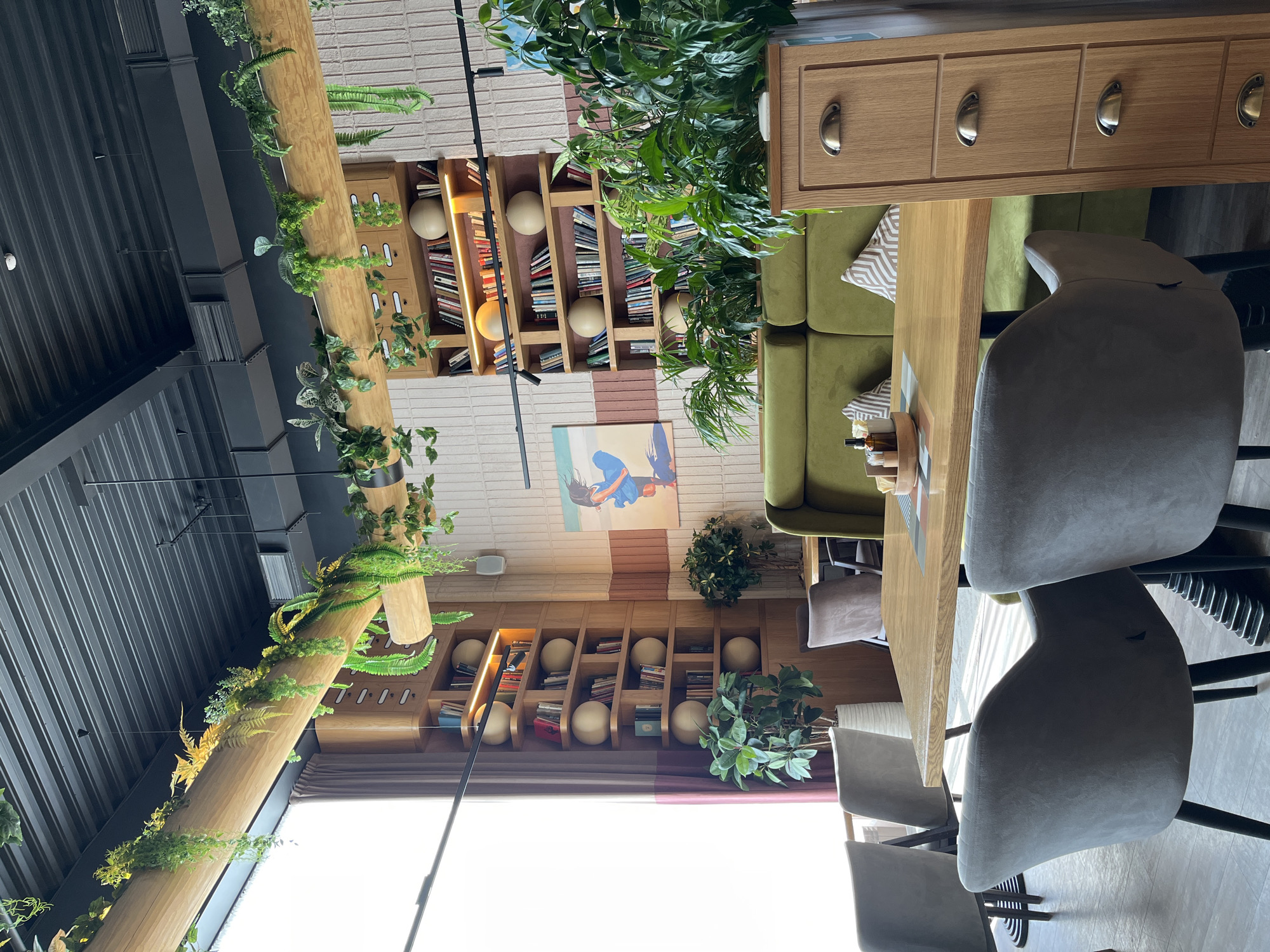 The image captures an indoor scene in what appears to be a restaurant or cafe in Verkhnyaya Pyshma, Russia. The viewpoint is from above, looking down at a table setting, chairs, and the surrounding decor.
The foreground features a wooden table with a tabletop pattern. Around the table are chairs upholstered in grey fabric with black legs. A green upholstered sofa extends behind the table.
Behind the seating area are bookshelves built into the walls. The bookshelves are filled with books, interspersed with large spherical decorative objects. Between the two sets of bookshelves is a painting or print on the wall.
Above, a wooden beam is suspended from the ceiling, adorned with hanging plants. A black metal structure, part of the building's framework, is also visible above.
The overall impression is of a space with a modern design, integrating natural elements like plants to create a comfortable atmosphere.