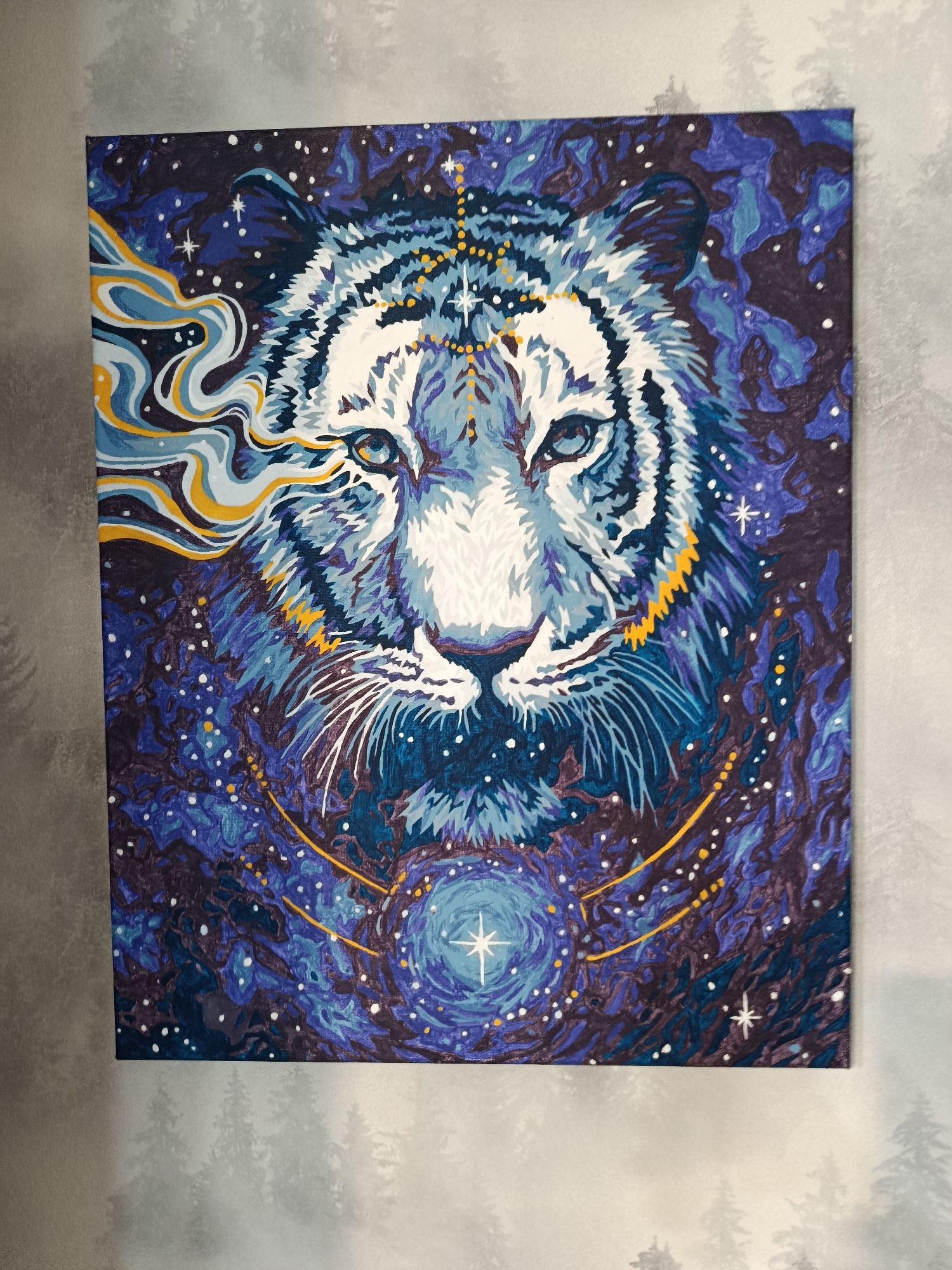 This is a close-up, eye-level, medium shot of a vibrant painting of a white tiger's face, rendered in a stylized, cosmic style against a deep blue and purple starry background. The tiger's fur is depicted with sharp, angular brushstrokes in shades of white, light blue, and dark blue, with yellow accents around its eyes and ears. Its piercing blue eyes are the focal point, surrounded by intricate facial markings. Swirling yellow and white smoke-like patterns emerge from the left side of the tiger's head, contributing to the ethereal feel. Below the tiger's face, a circular celestial motif is present, featuring a bright star at its center, surrounded by concentric blue rings. The painting is hung on a textured grey wall that has a subtle pattern of faint tree silhouettes. No specific location in the city of Apatity, Russia, can be identified from the image.
