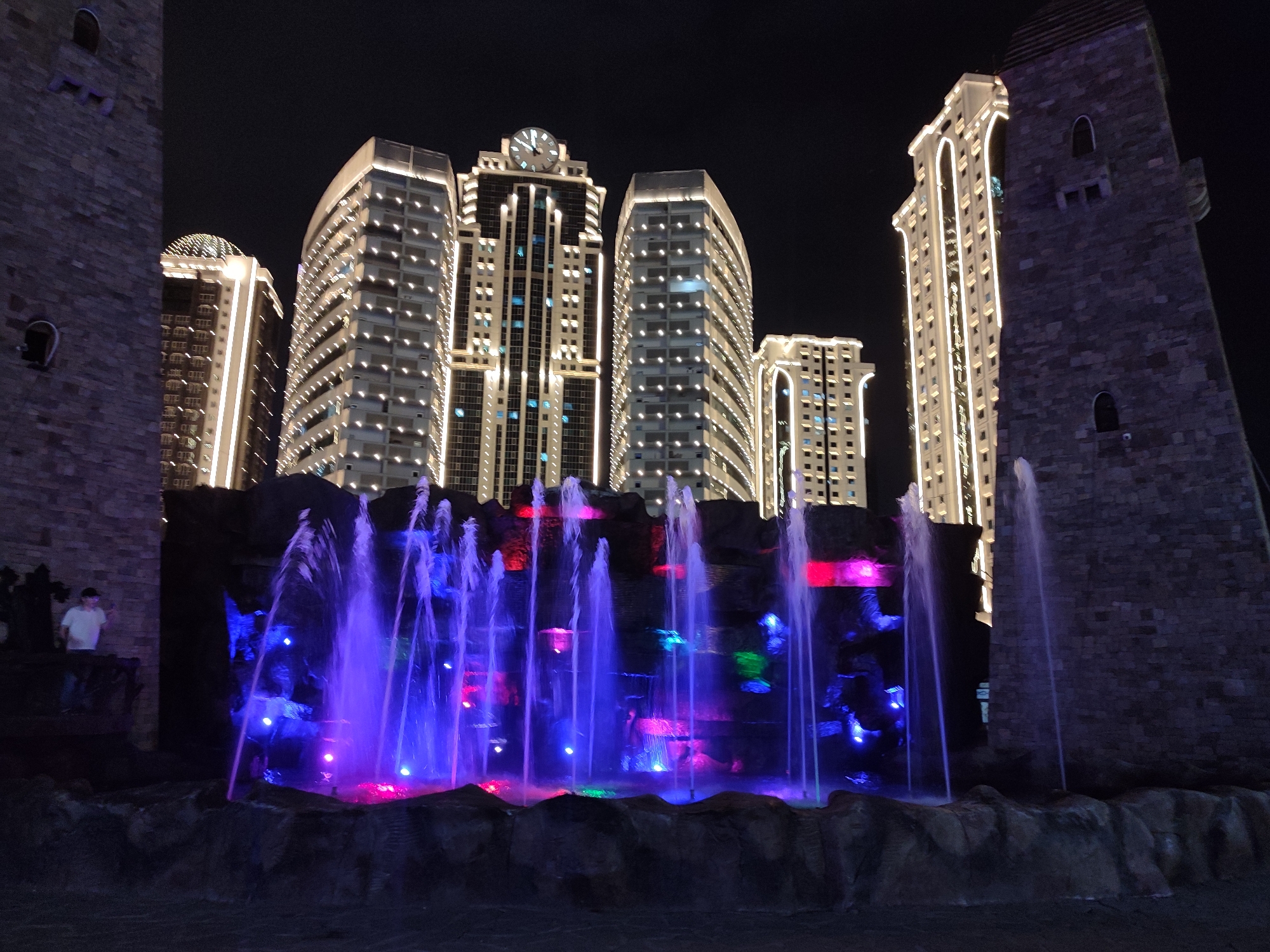 The image depicts a nighttime scene in Grozny, Russia, featuring a prominent water fountain illuminated with vibrant purple and pink lights. The fountain is set against a backdrop of modern, brightly lit skyscrapers, which stand out against the dark night sky. On either side of the fountain, there are large, dark, stone structures that resemble castle towers, adding a unique architectural contrast to the scene. One of these stone structures, on the left side of the image, has small, dark openings that appear to be windows. A person wearing a white shirt and dark pants is visible standing on a ledge near the base of the left stone tower, looking towards the fountain. The overall impression is one of a modern urban landscape enhanced by dramatic lighting and a whimsical fountain design.