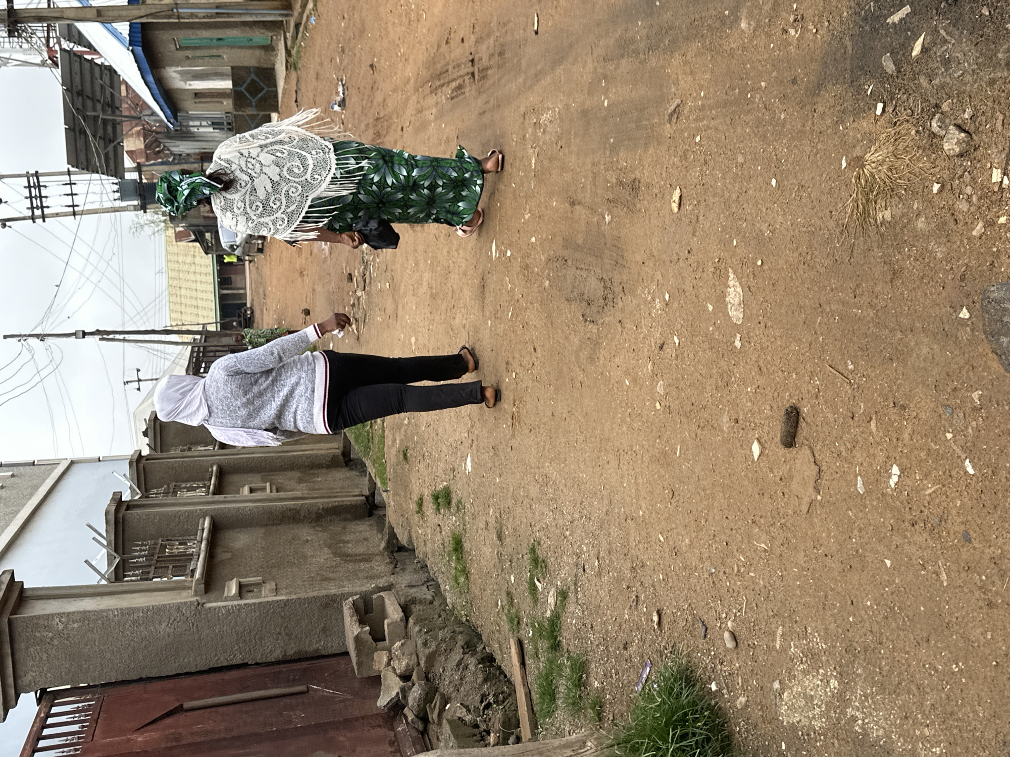 The image depicts a street scene in Narayi, Nigeria. Two women are walking away from the camera down a dirt road. The woman on the left wears a white hijab and a grey sweater with black pants. The woman on the right wears a green and white patterned dress and a white lace shawl. Both are wearing sandals. The road is lined with buildings and utility poles. There is debris scattered on the ground, including plastic wrappers and stones. The sky is overcast.