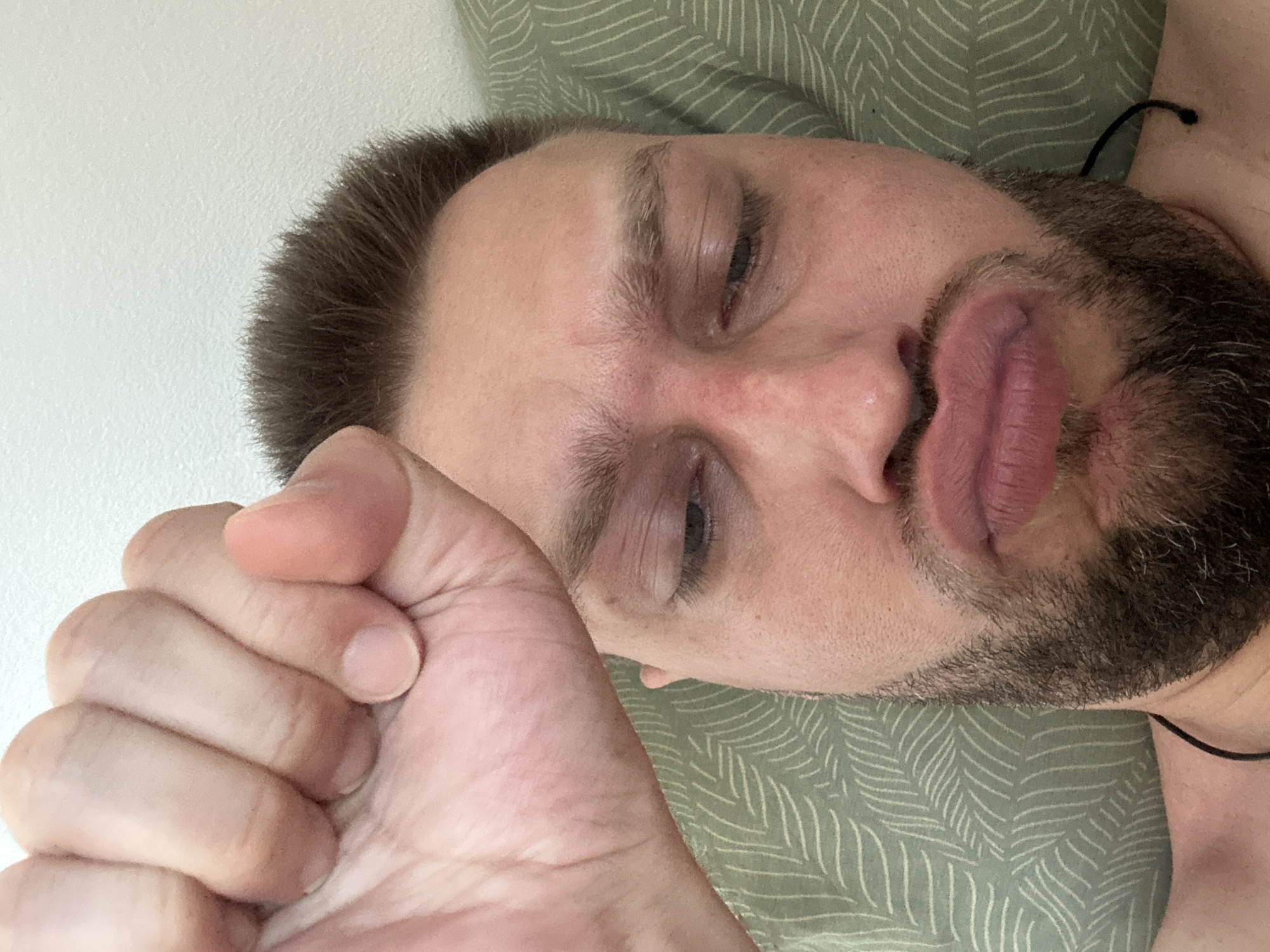 In this close-up, low-angle selfie, a fair-skinned man with brown hair and a beard lies on a light green pillow with a subtle leaf pattern. His right fist is raised and blurred in the foreground, obscuring a portion of his face. His left eye is visible, looking downwards, with an area of redness around his eye and on his cheek. His lips are pursed, creating a pout. The background consists of a plain, white wall. The man is wearing a thin black cord around his neck. The scene appears to be indoors, likely in a domestic setting, and does not provide specific indicators of a location within Zvezdara Urban Municipality, Serbia, beyond the possibility of it being a residential area.