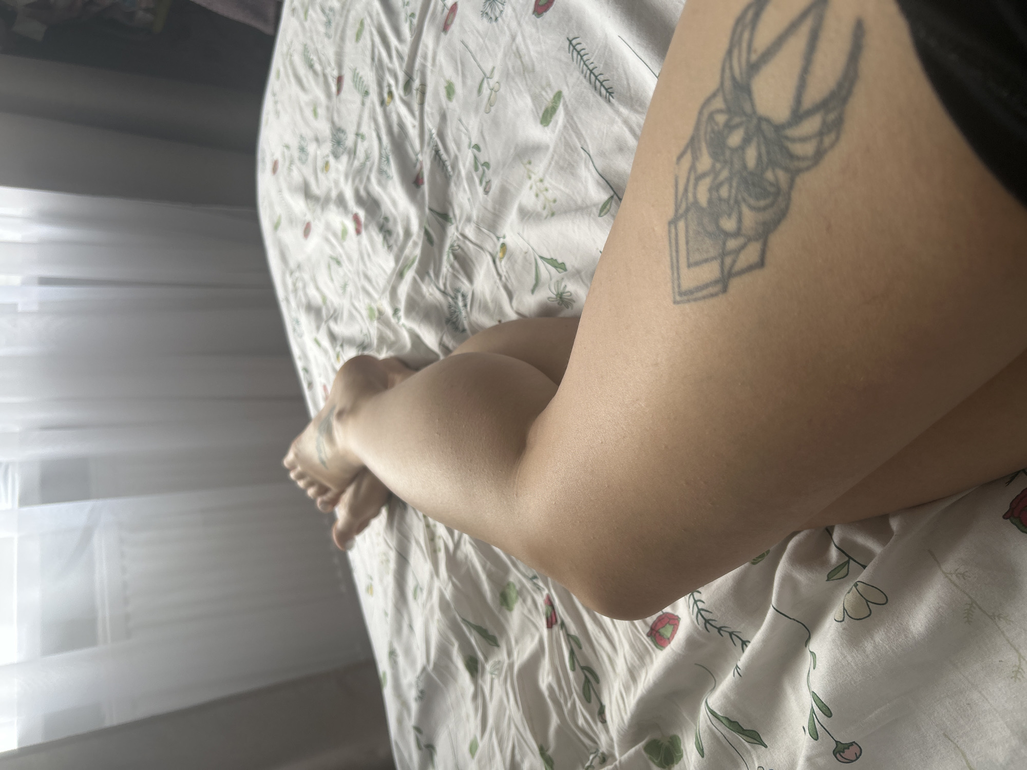 A first-person perspective shot captures a person's legs, crossed at the knees, resting on a bed with a floral-patterned comforter. The legs are tanned and bare, with a tattoo visible on the right thigh. The background shows a window with sheer curtains and a radiator, suggesting an indoor setting, possibly a bedroom. There is no indication of the image being located in Saint Petersburg, Russia.
