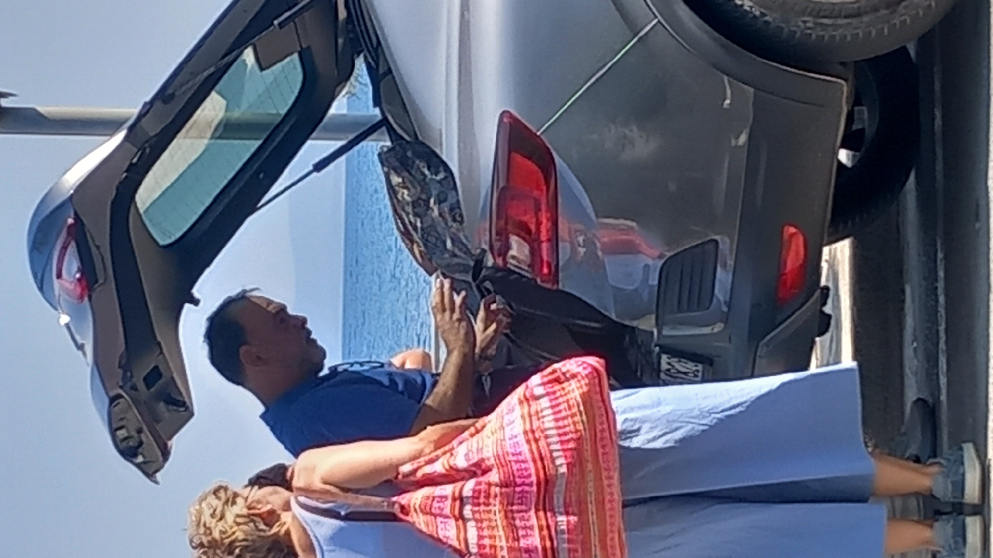 In Kos, Greece, a man in a blue shirt is seen with his hands inside the open trunk of a gray car. To his left, a woman wearing a light blue jumpsuit and carrying a pink and orange patterned tote bag stands beside the car. The scene appears to be outdoors, with the sea and a clear blue sky in the background, suggesting a location near the coast. The man's focus is on the contents of the trunk, while the woman stands passively, holding her bag. The overall impression is a moment of packing or unpacking luggage.
