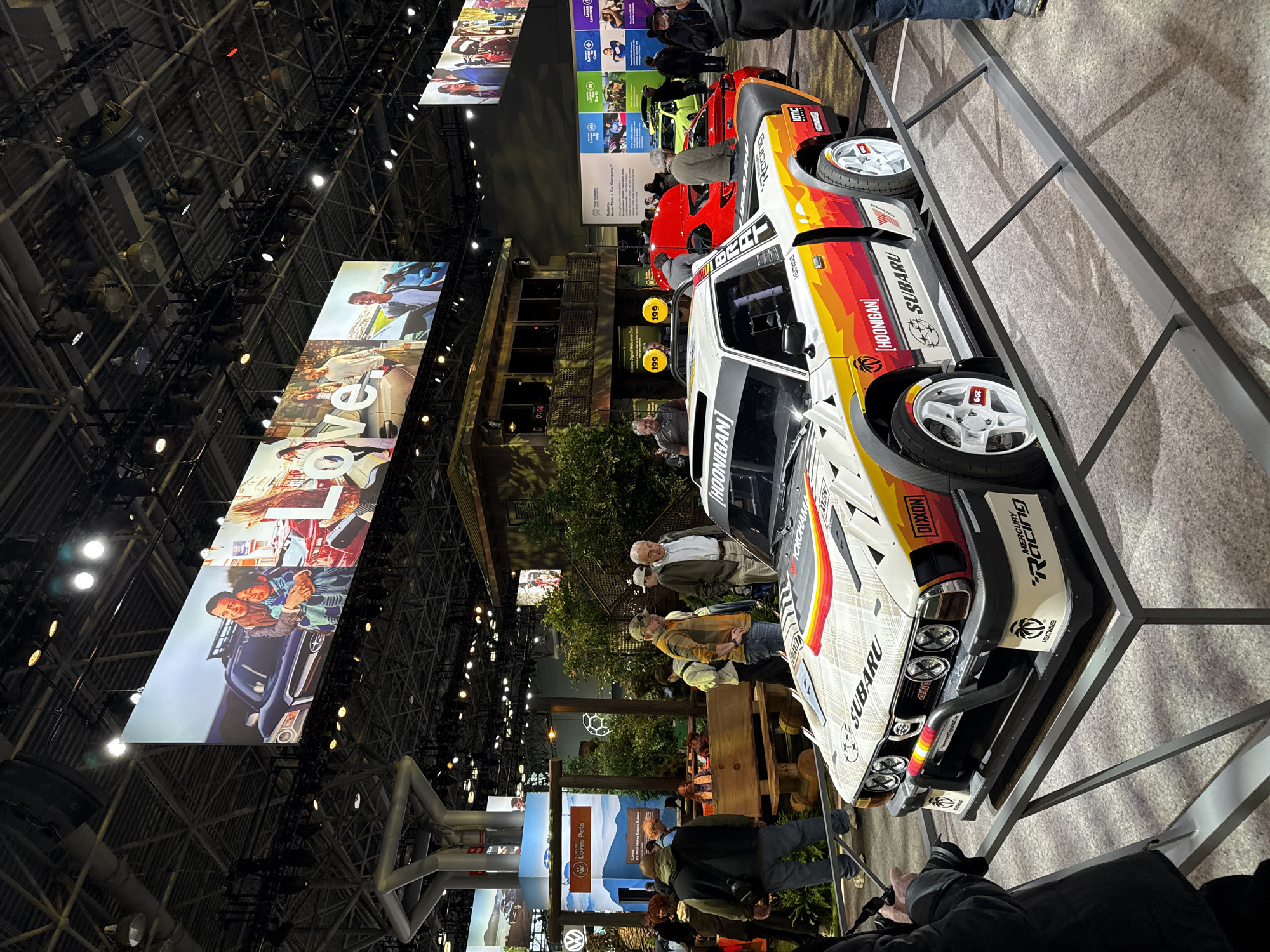 The image shows an indoor setting, possibly a car show or exhibition. A white Subaru BRAT race car with a distinctive livery is prominently displayed on a raised platform. Various logos and decals, including "HOONIGAN" and "SUBARU" can be seen on the car. People stand around the display, presumably observing the vehicle. A large screen hangs overhead, displaying a Subaru advertisement. The building's architecture includes a high, exposed ceiling with complex metal structures and lighting fixtures. The scene is brightly lit, highlighting the exhibits and attracting attention.