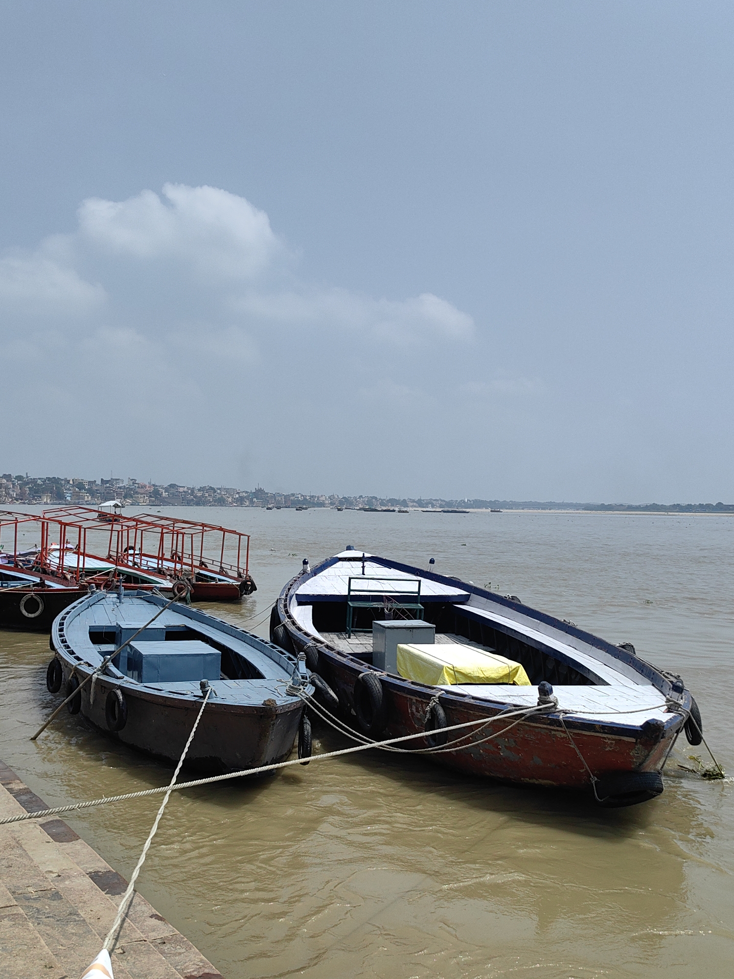 The image shows a view of the Ganges River in Varanasi, India, with several boats docked along the riverbank. In the foreground, a blue boat is tied to the shore, and to its right, a larger boat with a red and white hull is also moored. Behind these boats, a row of smaller boats with red canopies is visible. The cityscape of Varanasi is visible in the distance across the river, characterized by its numerous buildings and temples. The sky is overcast with some clouds. The water of the Ganges appears muddy.