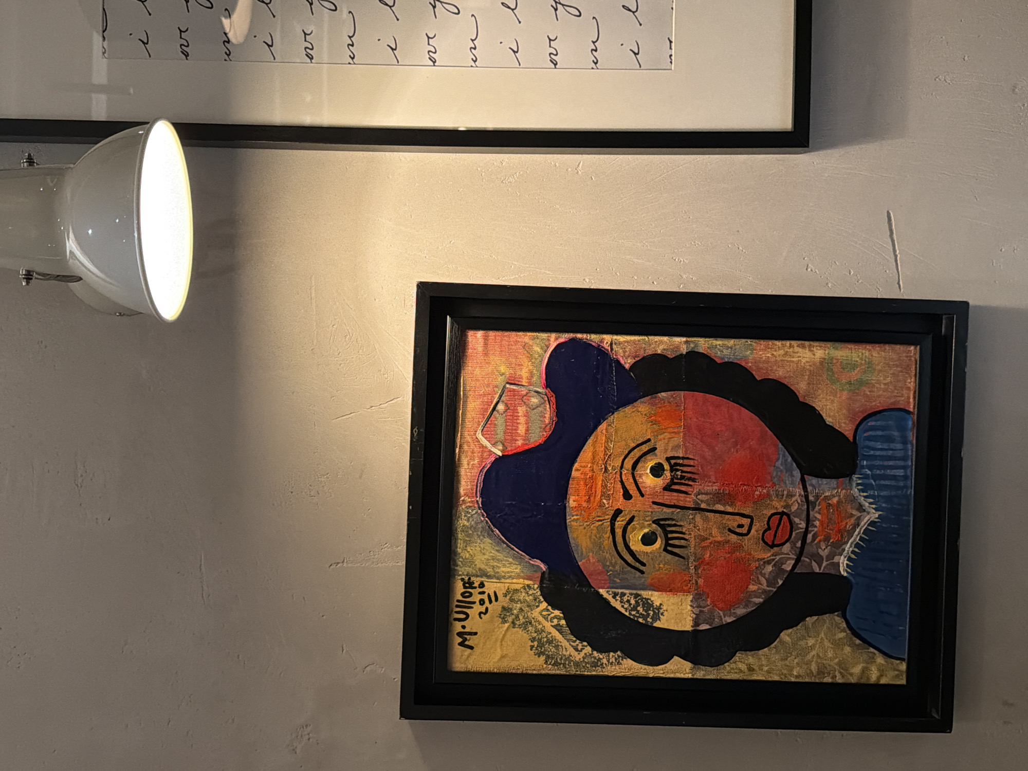 The image shows a wall with two framed artworks and a lamp. The top artwork features script in a white frame with a black border. Below it, to the right, is a smaller, abstract painting in a black frame. The painting depicts a face with stylized features and bold colors like red, blue, yellow, and black. A white lamp is mounted to the left of the upper artwork, shining its light onto the scene. The wall appears to be textured and painted in a light beige or gray color. The lower artwork is signed "M-Ullop 2011".