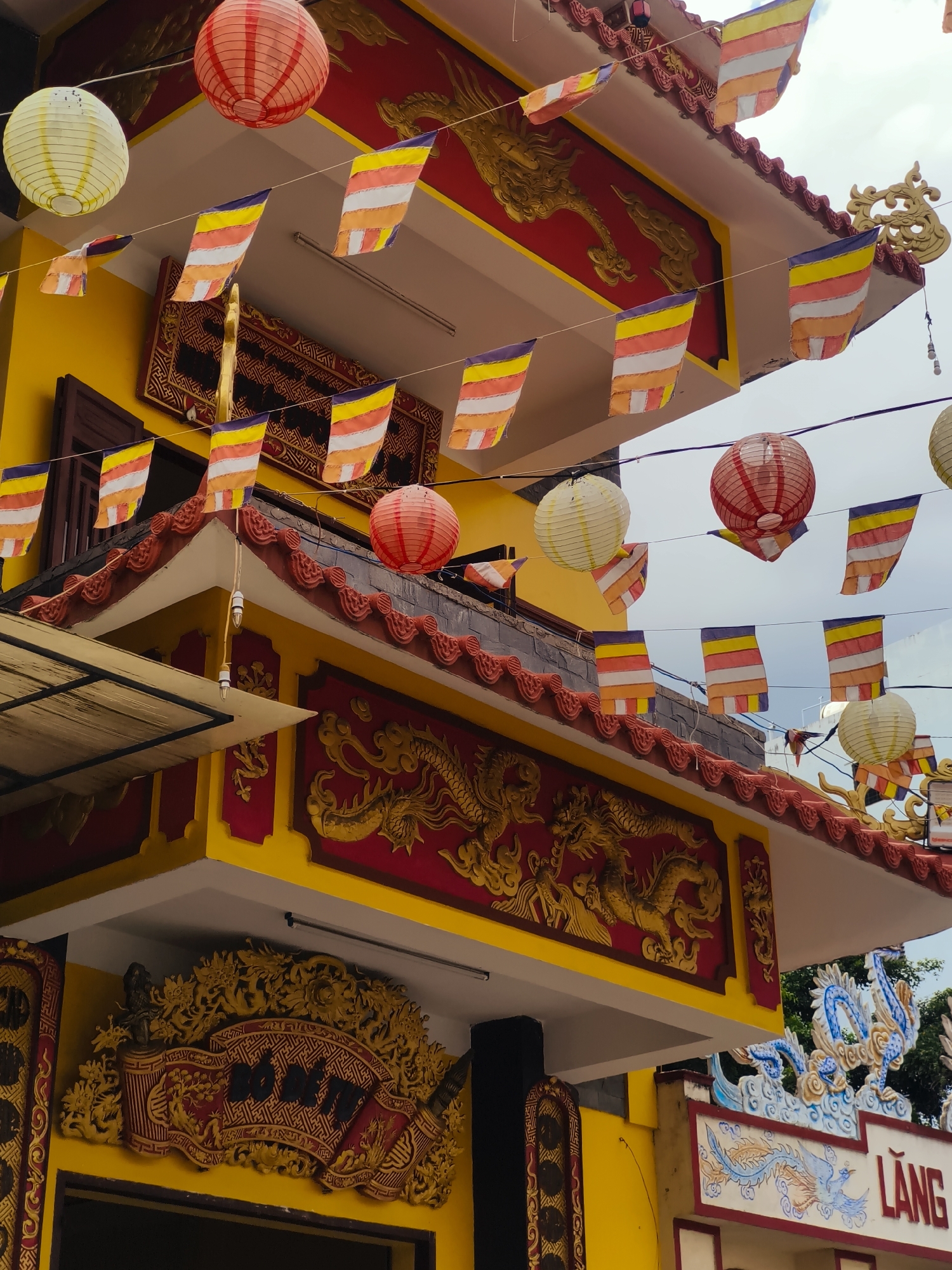 The image captures an exterior view of a vibrant yellow building, likely a temple or cultural structure, in Nha Trang, Vietnam. The architecture features a red-tiled roof with decorative elements and ornate gold carvings of dragons adorning the facade. Strings of colorful prayer flags, with alternating stripes of yellow, orange, white, and purple, are strung across the scene, fluttering in the breeze. Several paper lanterns, in shades of red and white, are also suspended, adding to the festive atmosphere. The lower portion of the building displays a large, intricate gold relief on a red background, depicting a dragon in motion. To the right, another section of the building is visible with blue and white dragon motifs and the partial inscription "LÀNG" in white letters on a red background, suggesting it might be part of a name or signage. The sky above is overcast with a pale blue hue. There are no people visible in the frame. The overall impression is one of traditional Vietnamese religious or cultural decoration.