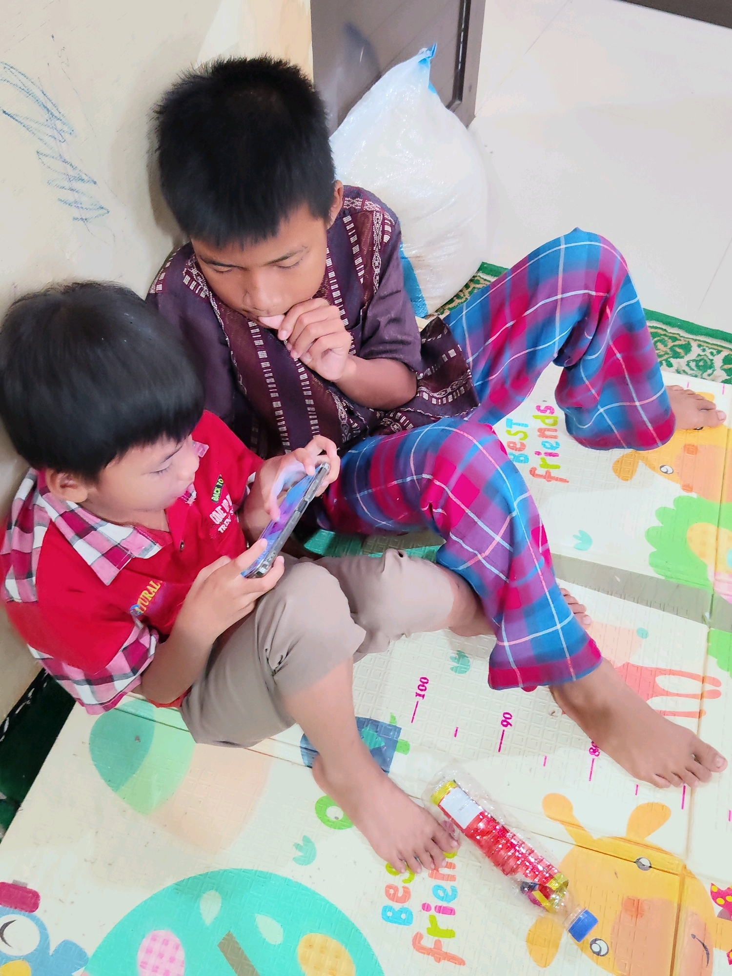 The image depicts two young boys sitting on a soft play mat in what appears to be a domestic indoor setting. The boy on the left, wearing a red collared shirt with a plaid pattern on the collar and sleeves, is holding a smartphone and appears to be playing a game. He has short, dark hair and is wearing tan shorts. The boy on the right, wearing a dark patterned shirt and plaid pants with pink and blue squares, is looking down and has his finger near his mouth, possibly thinking or biting his fingernail. He also has short, dark hair. Their legs are crossed, and they are seated closely together.
The play mat they are on has a colorful design with various shapes and what looks like measurement markings. A green and white patterned rug is visible underneath the play mat. In the background, a large white bag, possibly filled with something soft like cotton or plastic, is propped against a wall. The wall to the left has some faint blue scribbles or drawings on it. A clear plastic bottle with red and yellow detailing, containing small colorful objects, lies on the play mat near the feet of the boy on the right. The overall lighting suggests it is daytime, likely indoors. The presence of a play mat and the casual seating arrangement suggest a home environment, possibly a living room or playroom.
While the image does not provide explicit clues to pinpoint the exact location within Purwakarta, Indonesia, the general scene is consistent with a typical Indonesian household. The clothing styles of the children, the presence of what could be a prayer mat, and the commonality of soft play mats for children all align with cultural practices and material goods found in Indonesia. The image captures a moment of shared activity, with both children focused on their own engagement, though in close proximity.