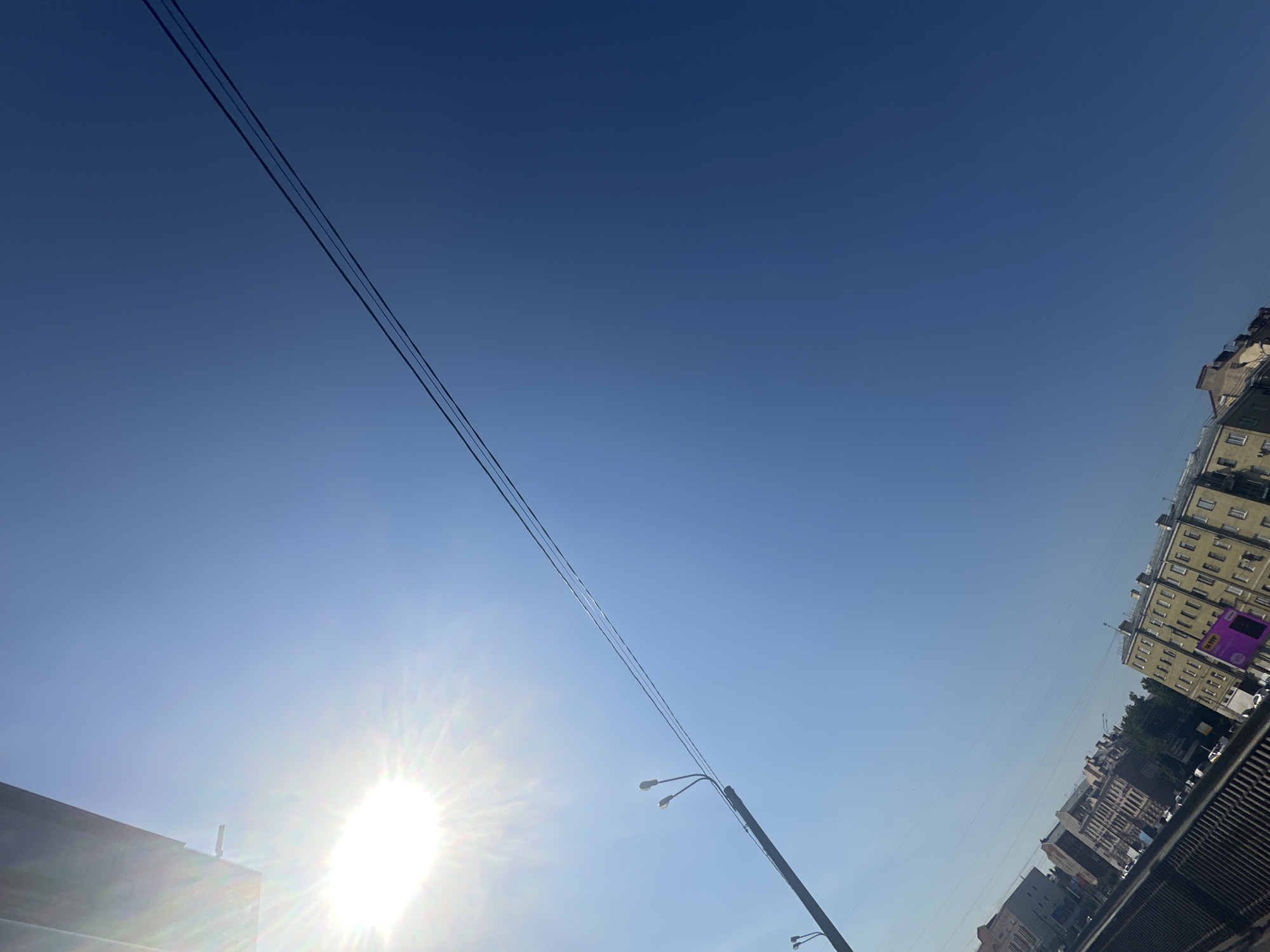 The image captures a bright, sunny day in an urban environment, likely in a city. The sky is a clear, deep blue, with the sun prominently positioned in the upper left quadrant, emitting a strong lens flare. Several parallel wires stretch diagonally across the sky from the middle left to the upper right. In the lower portion of the frame, the rooftops and upper facades of buildings are visible, suggesting a street-level or elevated perspective. The architecture appears to be of a European style, with multi-story buildings featuring numerous windows. A street light with two lamps is attached to a pole on the left side of the image. The overall scene suggests a typical urban landscape under a clear sky. The specific location is not identifiable from the visual information provided.