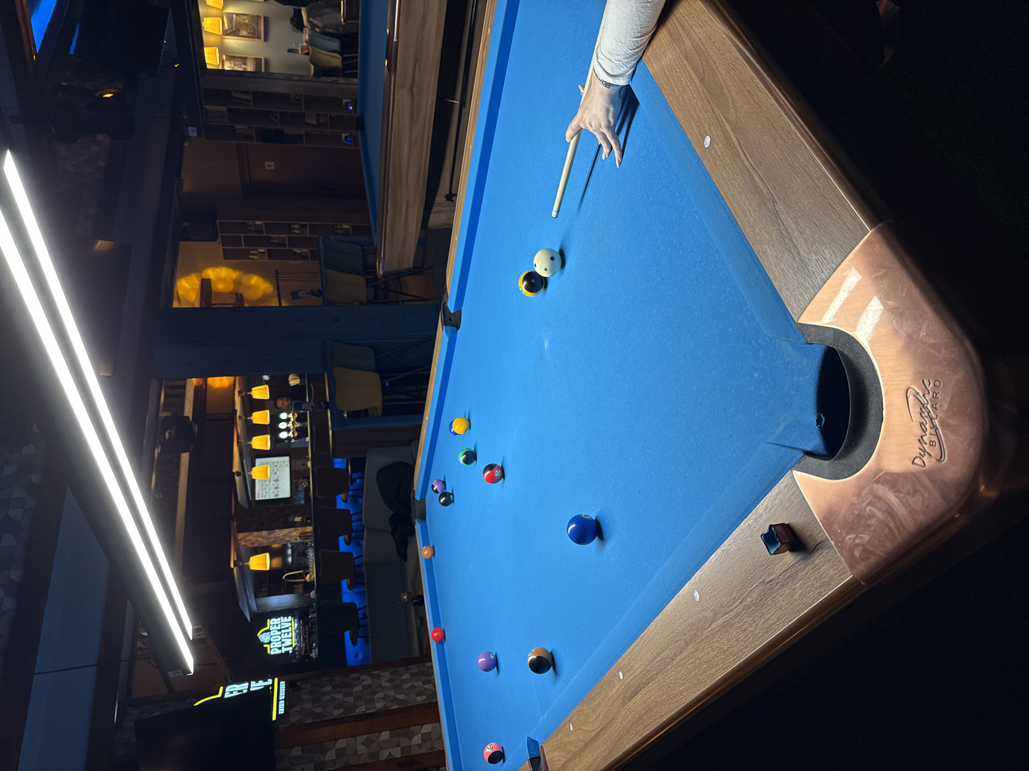 A slightly rotated and angled shot depicts a billiards table in what appears to be a bar or entertainment venue. The table has a blue felt surface with numerous billiards balls scattered across it. A person’s hand and cue stick are visible near the top right corner of the table, implying a shot is about to be taken.
The background shows an interior space with lighting fixtures, seating, and a bar area. The style of the bar resembles a modern American or European bar.
Without additional information, the location cannot be specified in detail to Plovdiv, Bulgaria.