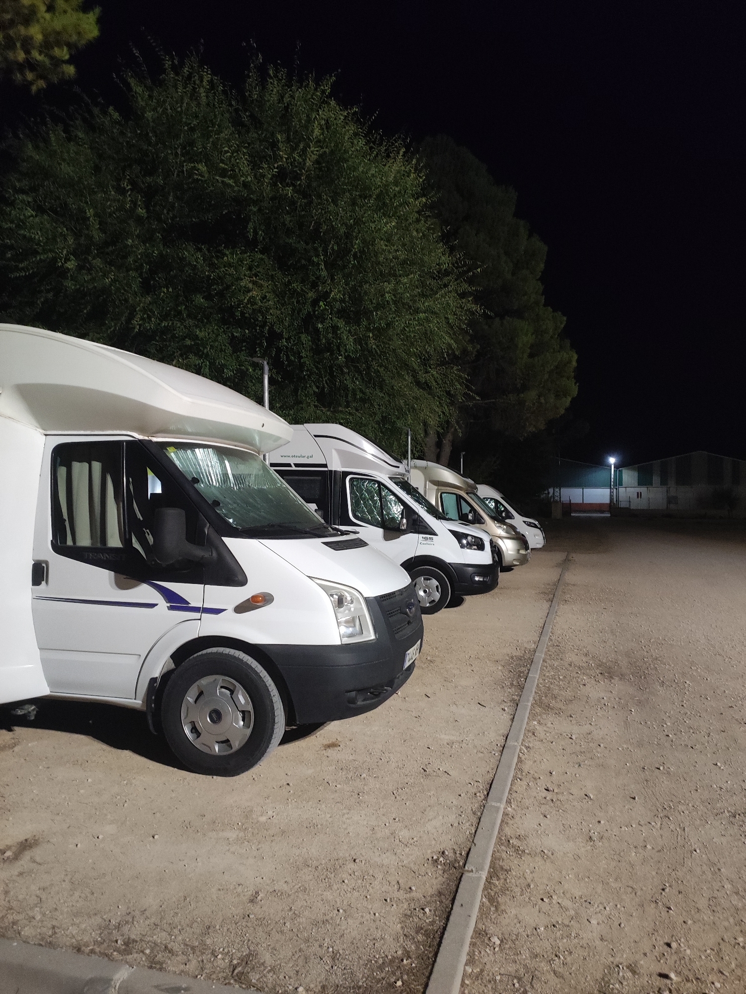 In the city of Villarejo de Fuentes, Spain, at nighttime, a row of white recreational vehicles (RVs) is parked side-by-side on a gravel lot adjacent to a line of trees. The RVs are facing right, with the closest one, a white Ford Transit, prominently featured on the left side of the frame. It has its driver's side door open. The RVs are illuminated by a single streetlight in the background, casting shadows on the ground. In the distance, a building with a white facade and a green roof is visible. The overall scene is calm and quiet, typical of a campsite or parking area at night.