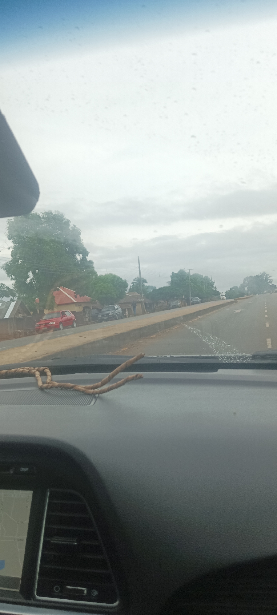 The image is taken from inside a vehicle, likely a car, looking out onto a street scene in Gwadanye, Nigeria. The dashboard of the car is visible, with a knotted brown rope and part of the navigation screen. The windshield has water droplets on it.
The street outside is asphalt and features a curb or sidewalk dividing it from the buildings and foliage. A red car and other parked cars are visible along the side of the road, near what appears to be a residential or commercial area with trees and buildings. The sky is overcast.