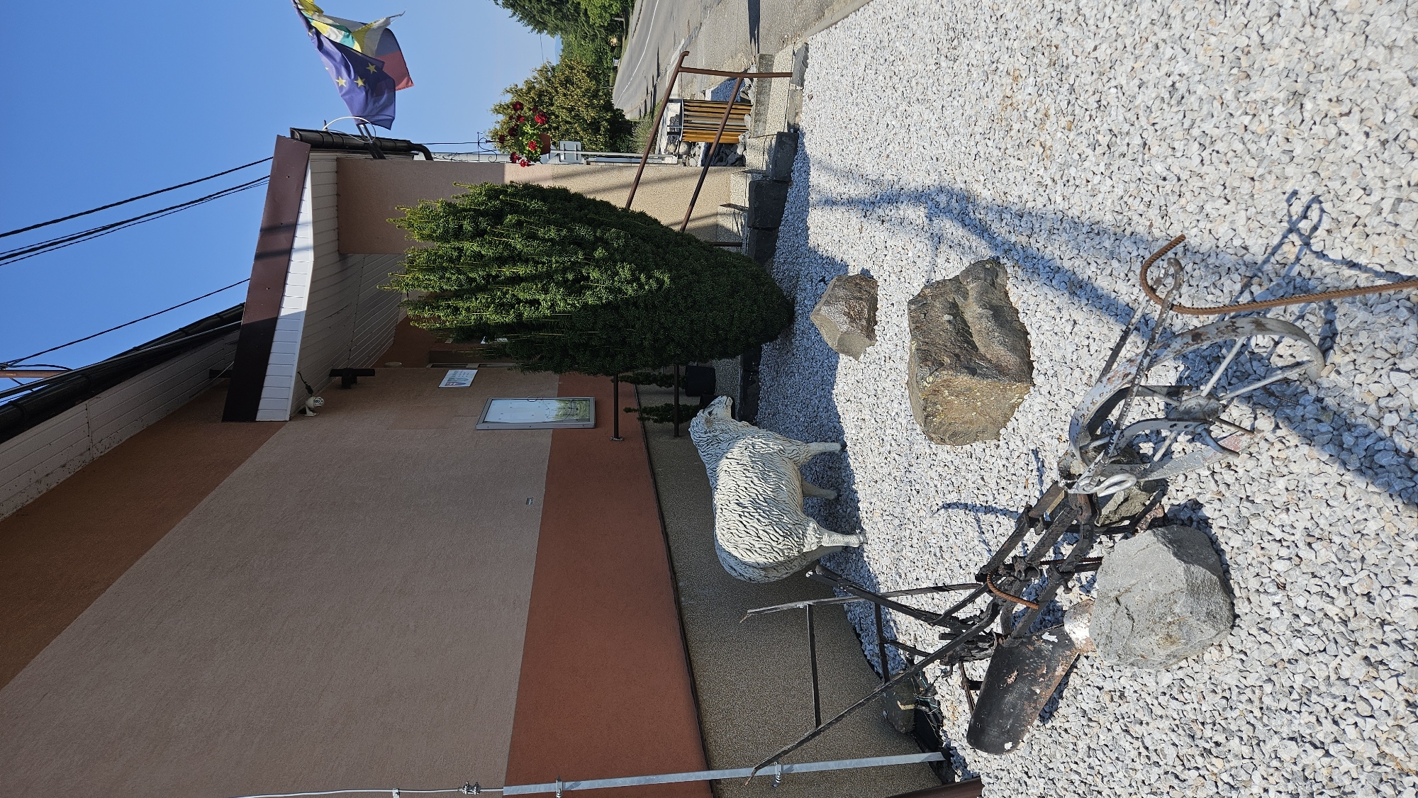 In the city of Vyšné Nemecké, Slovakia, a white, weathered sheep statue stands in a gravel-covered yard next to a building with a tan and orange facade. A large, dark green, conical evergreen tree is situated behind the sheep statue, partially obscuring the building. In front of the sheep, a collection of large, gray rocks are scattered across the gravel. To the left of the sheep, a rusted, old farm implement, possibly a plow, lies on the ground, also surrounded by gravel and a few rocks. A security camera is mounted on the building above the entrance. The scene is illuminated by bright sunlight, casting distinct shadows. A paved road and more greenery are visible in the background, under a clear blue sky. The EU flag is flying from a pole attached to the building.