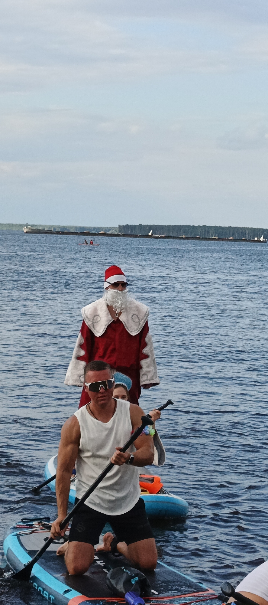 The image captures a group of people on paddleboards in a body of water, likely a river, under a cloudy sky. The water is blue with moderate ripples, suggesting a breezy day. In the foreground, a man wearing a white tank top and black shorts is kneeling on his paddleboard, holding a paddle. He is wearing dark sunglasses. Behind him, a person dressed as Santa Claus stands on a paddleboard, complete with a red suit, white fur trim, a red hat, and a white beard. Another person, partially visible, is seated behind Santa. In the background, a long barge is visible on the water, with a line of trees on the horizon. Further in the distance, smaller boats and what appear to be sailboats are scattered across the water. The overall scene suggests a recreational activity or event, possibly a themed paddleboarding outing. The lighting is diffused due to the overcast sky, indicating it's daytime. The text "SARNAT" is visible on the paddleboard in the foreground.