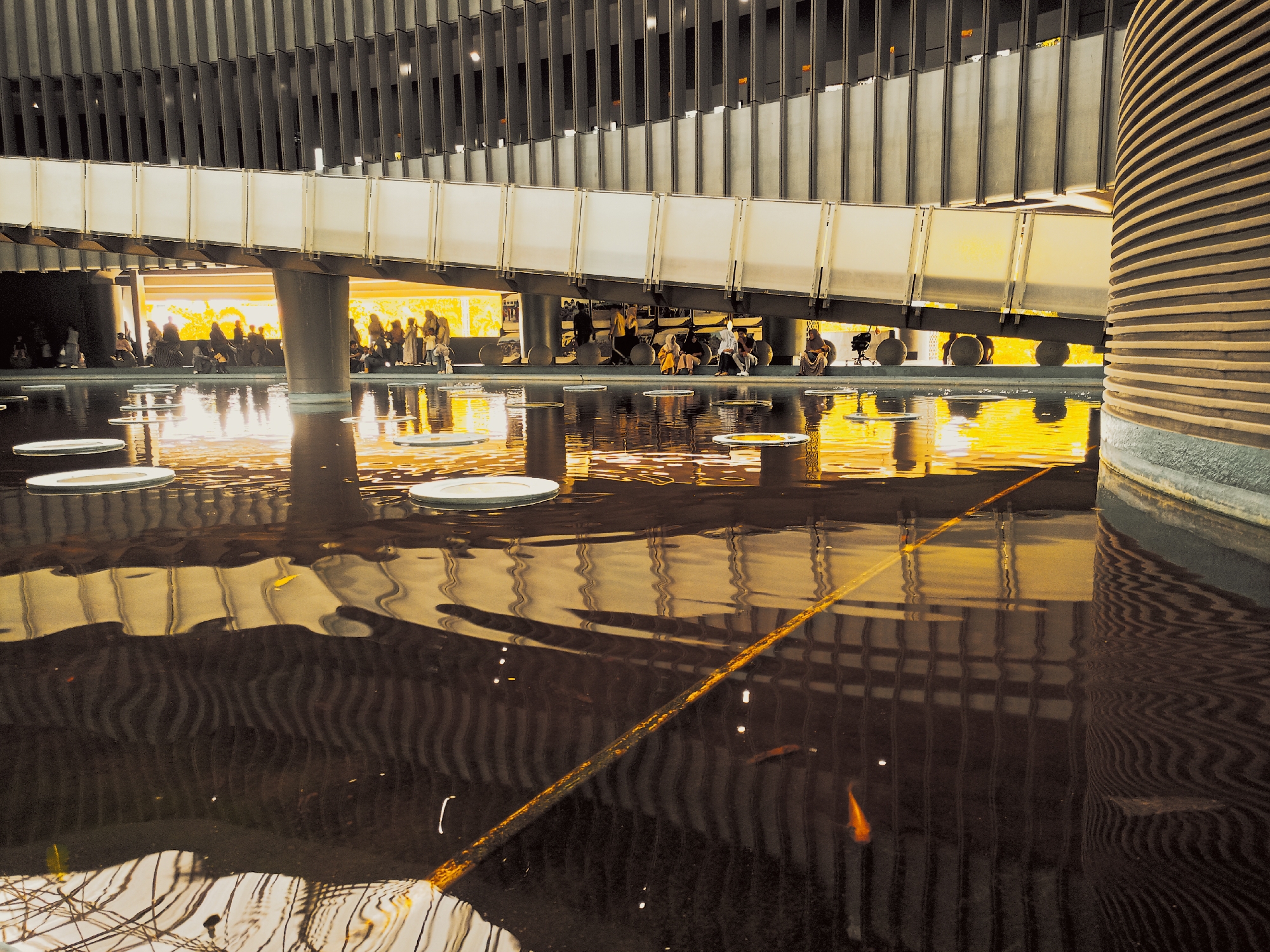 An eye-level medium shot shows a water feature in an exterior location, possibly in Banda Aceh, Indonesia. Reflected in the water are the architectural features of a curved, elevated walkway supported by columns, with vertical, parallel slats above the walkway. The walkway is illuminated from behind, and its light is strongly reflected in the water, creating a bright, amber-colored glow. People are indistinctly visible behind the walkway. White, floating disks are scattered across the water's surface. The water is mostly dark and rippled, with faint reflections of the walkway's structure and surrounding environment. A few orange fish are visible swimming in the water.