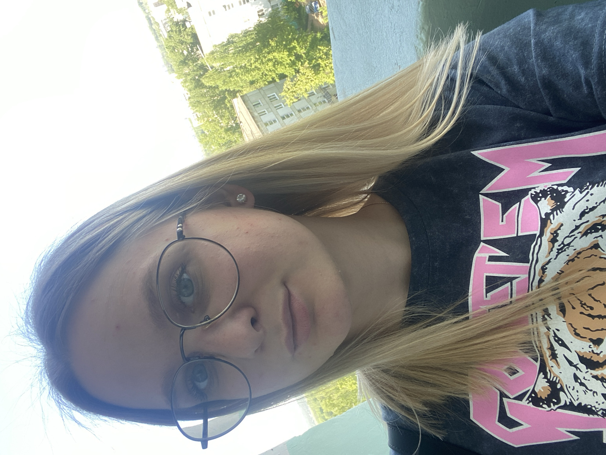 A selfie of a young woman with blonde hair wearing round glasses and a t-shirt with a pink and black tiger design. She is looking directly at the camera with a neutral expression. The background shows buildings and trees, suggesting an urban environment. The specific location and city are not discernible from the image.