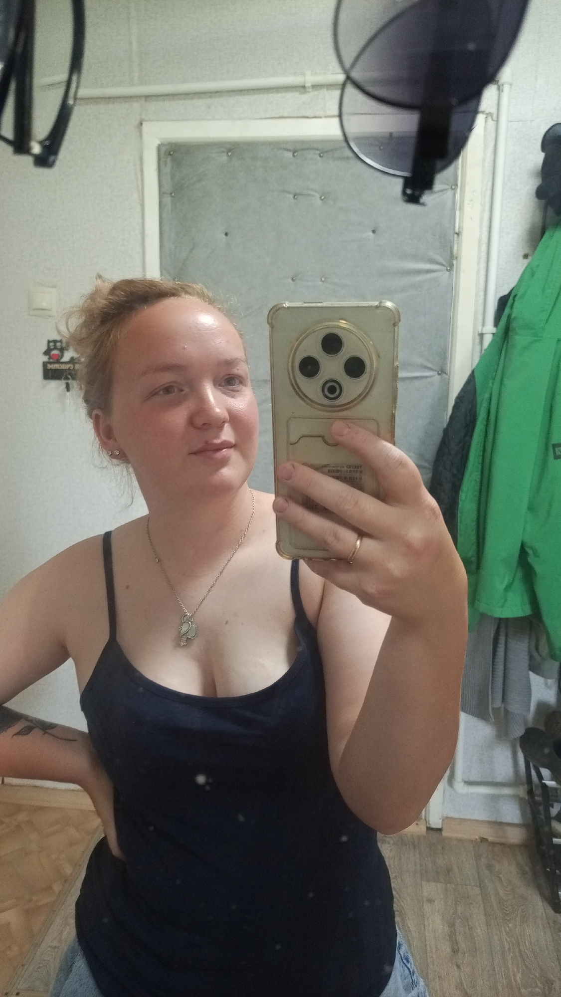 A woman with fair skin and light brown hair pulled back in a messy bun is taking a selfie in what appears to be a bathroom. She is wearing a dark blue tank top and a silver necklace with a pendant. A gold ring is visible on her ring finger. She holds a gold-colored smartphone with a distinctive triple-camera setup. The background includes a padded door, a green jacket hanging on a rack, and a hairdryer mounted on the wall. The location is assumed to be in Уральское, Russia, based on unspecified information.