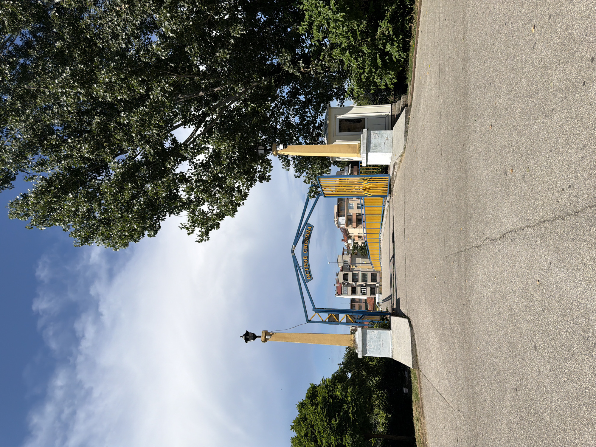 The image depicts the entrance to Freedom Island (Ostrov Svoboda) in Pazardzhik, Bulgaria. A paved pathway leads towards a bridge with a yellow and blue archway inscribed with "ОСТРОВ СВОБОДА" in yellow letters. Flanking the entrance are two tall, yellow, concrete pillars topped with black lamps. To the right of the right pillar is a small, white booth with a door. The pathway is bordered by concrete structures and lush green trees. In the background, a row of multi-story buildings is visible. The sky above is a bright blue with scattered white clouds. The overall scene is well-lit, suggesting it was taken during the daytime. There are no people visible in the frame.
