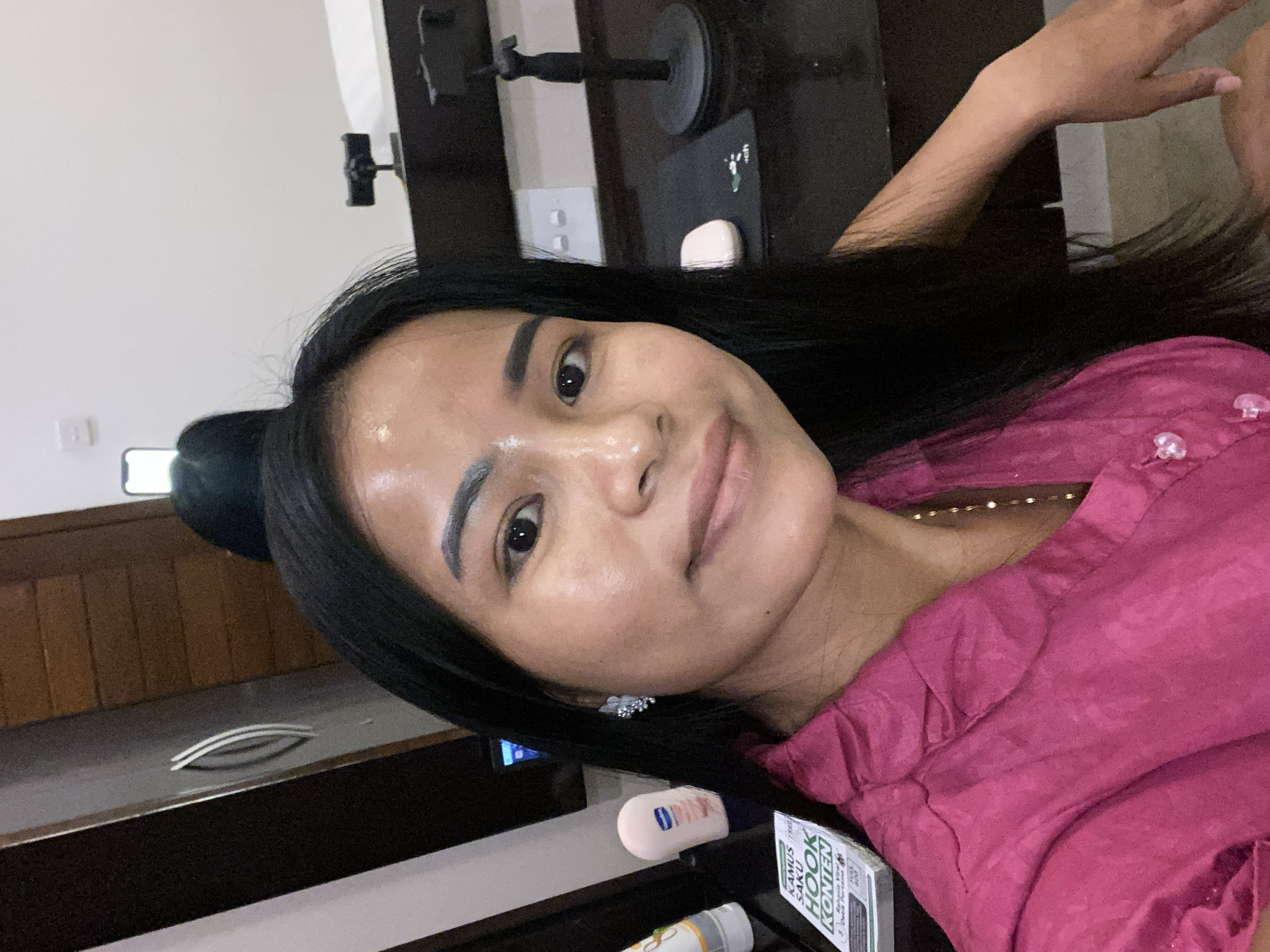 The image shows a woman indoors, likely in a bedroom, based on the presence of a drawer or cabinet unit. The woman has dark hair and is wearing a magenta-colored top. She is positioned in front of a dark-colored desk or surface where several items are placed, including what appears to be a bottle of lotion and a small book, "Kamus Hook Konien."
In the background, there is what looks like a phone holder and some other objects on the desk. The overall setting has a casual feel. There's no direct indication of the location being North Kuta, Indonesia from visual elements in the image. The location is based on the user's provided information in the prompt and has no relevance to the image itself.