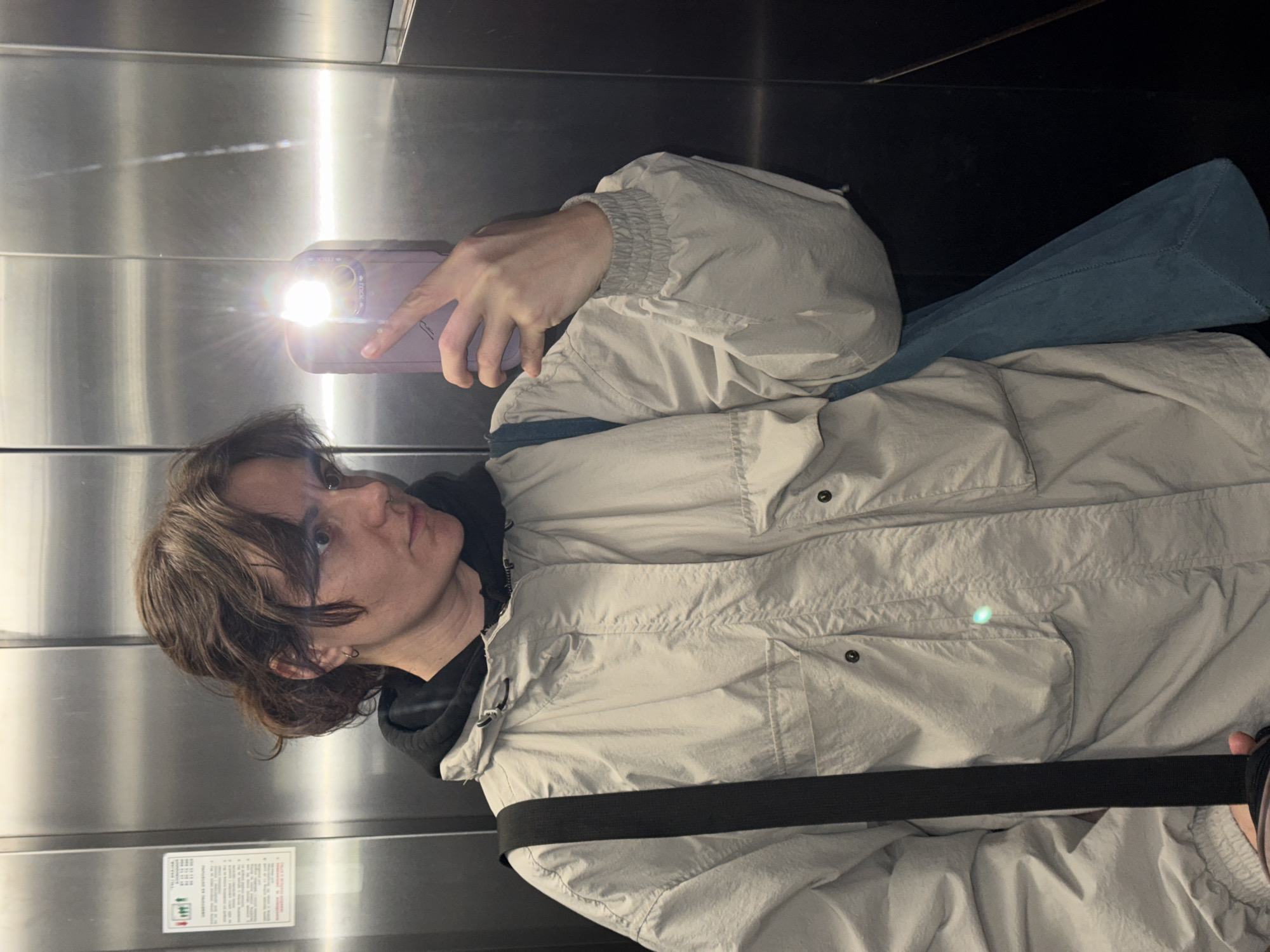 A person with brown hair is taking a selfie in a reflective elevator. The person is wearing a light-colored jacket over a dark hoodie, with a dark strap across their chest. The background is a metallic elevator interior. There's a flash of light from the phone's camera. The scene is not necessarily set in Boreti, Montenegro.