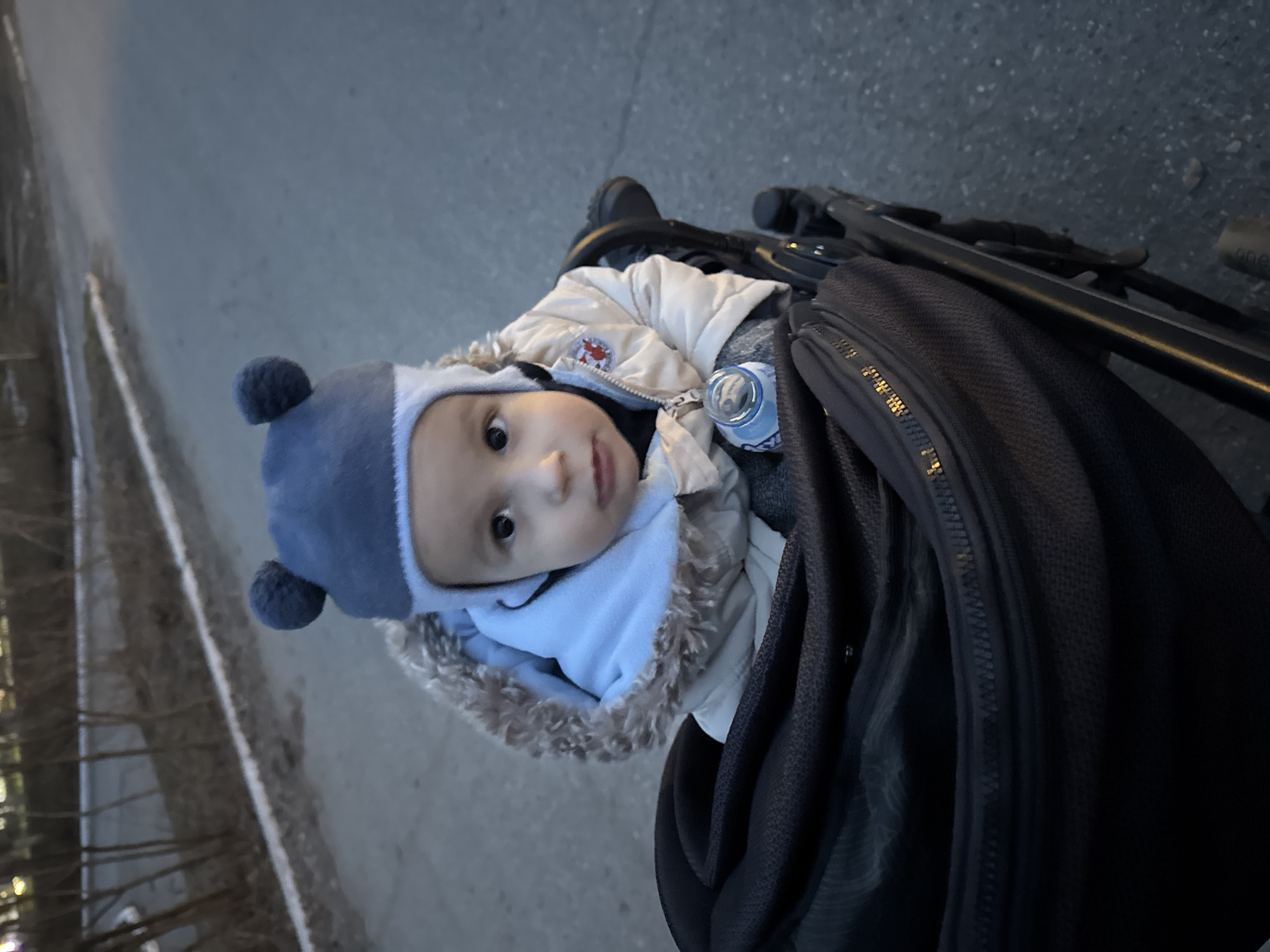 A baby wearing a blue hat with pom-poms and a fur-trimmed jacket is sitting in a stroller. A bottle of water is visible next to the baby. A black bag is attached to the stroller, and the stroller is on a paved surface. The location is possibly in the city of Chaikovsky, Russia. The lighting suggests it is daytime, potentially late afternoon or early evening.