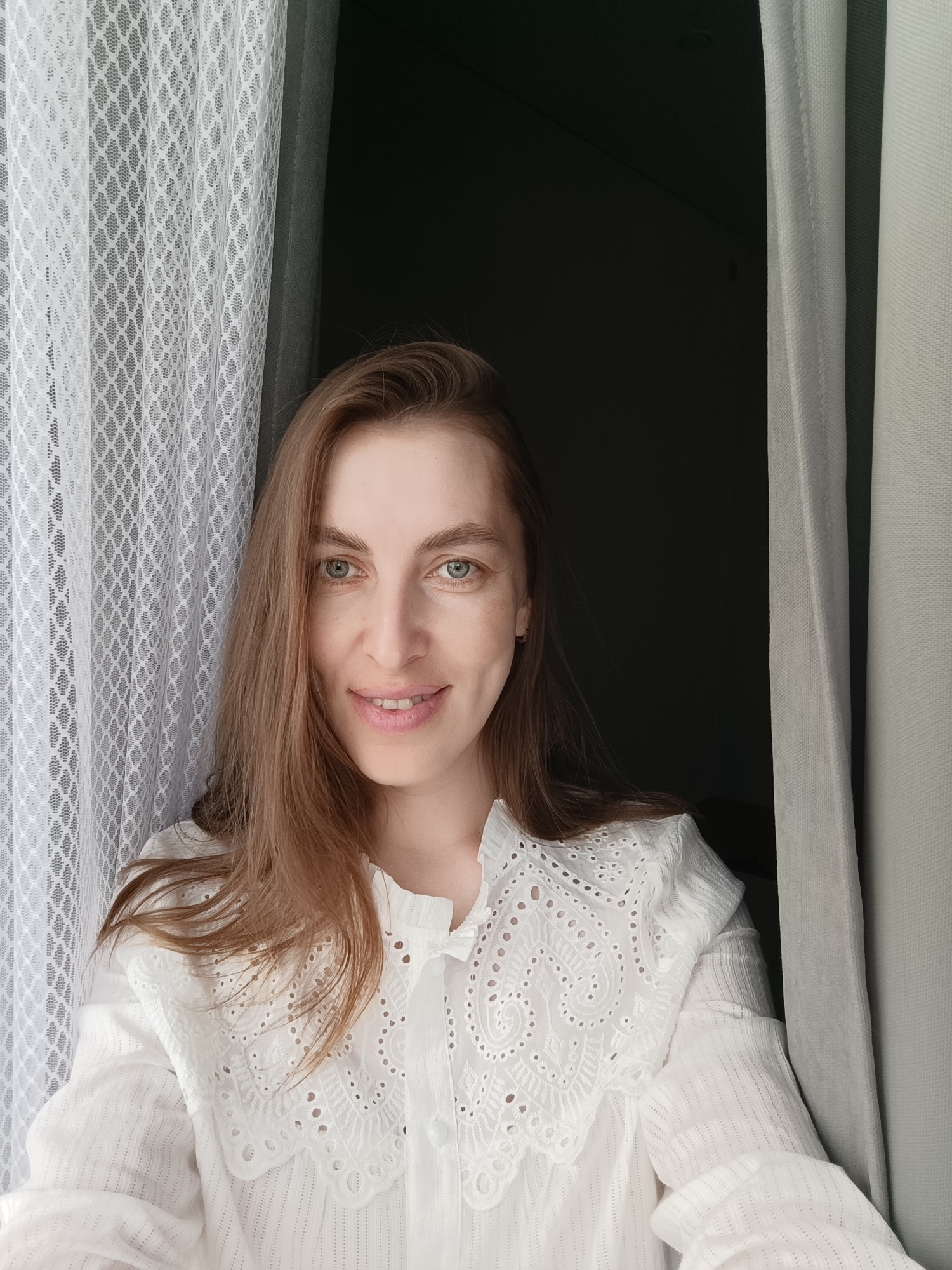 A Caucasian woman with long brown hair and green eyes is centered in a medium shot, appearing to be indoors. She is wearing a white blouse with lace detailing. She is positioned between two curtains; a white, lace curtain to her left and a solid gray curtain to her right, with a dark space visible between them. The location is Chaikovsky, Russia.