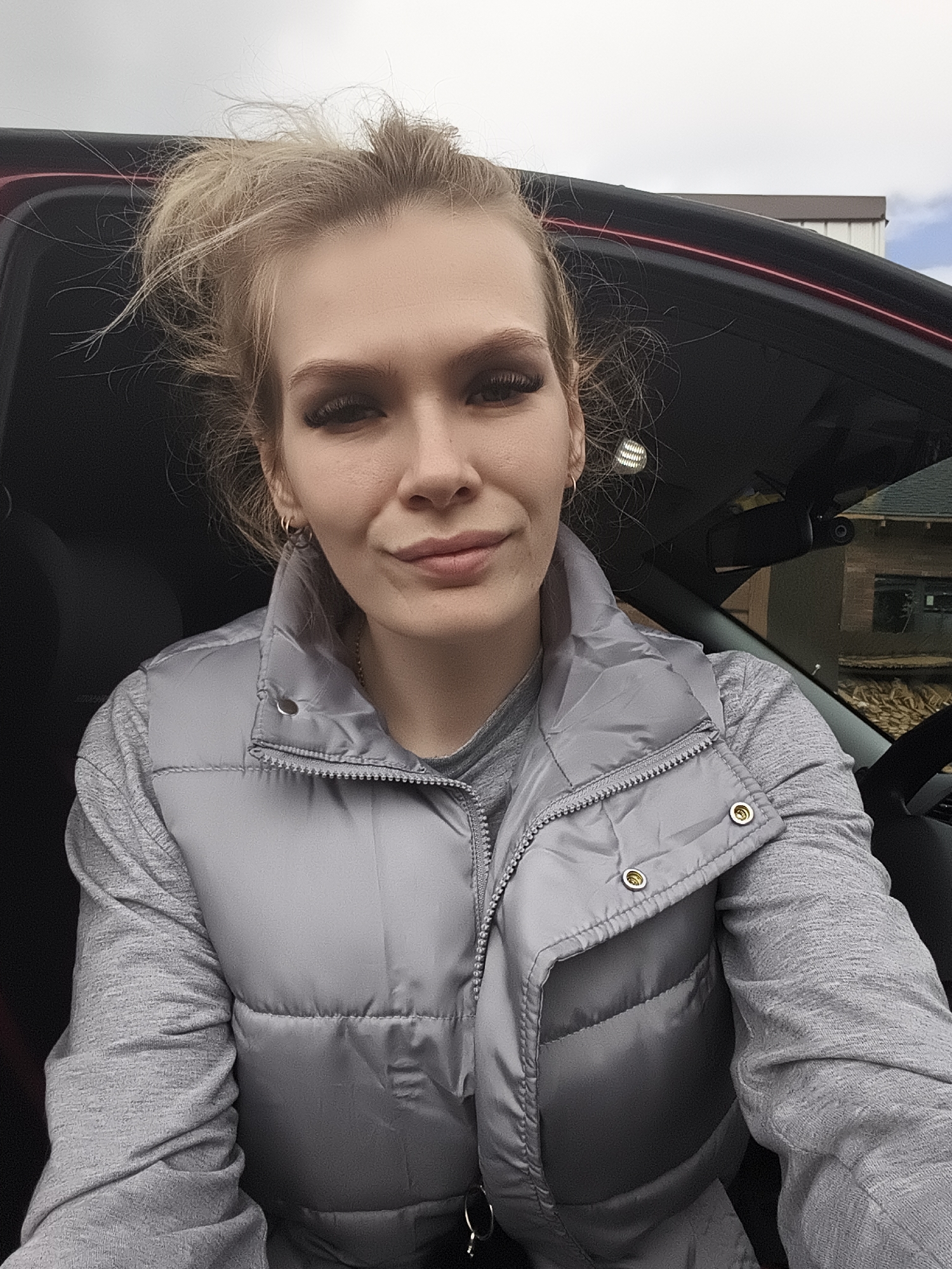 A woman sits in the driver's seat of a car. She has blonde hair pulled up into a messy bun and wears a gray padded vest over a gray long-sleeved shirt. Her car is parked in Русский Вожой, Russia. Behind the car, a brick building and a partly cloudy sky can be seen.