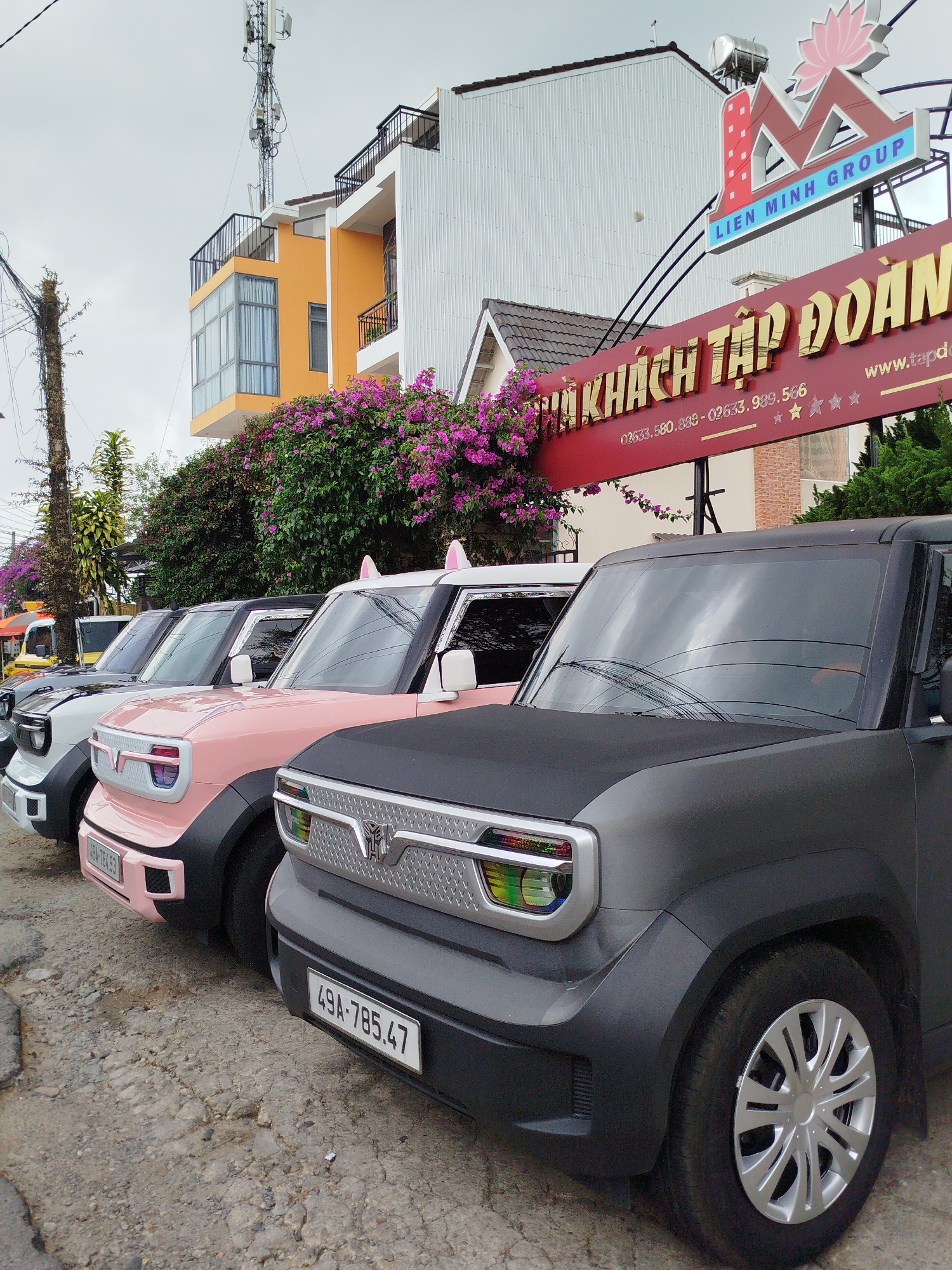 A row of distinctively styled electric vehicles (EVs) is parked on a rough, paved surface in front of a building in Da Lat, Vietnam, under an overcast sky. The cars are compact and boxy, resembling micro-EVs, and are presented in varying colors. The closest vehicle is matte dark gray, featuring a metallic silver grille with a Transformers logo and a rainbow-colored headlight surround. Its license plate reads "49A-785.47". Behind it is a pink EV with a white roof, adorned with pink cat-ear accessories, displaying the license plate "49A-784.93". Further back, another white EV with a black roof, and a dark-colored vehicle complete the line.
The background features a multi-story building, part of which is white with corrugated siding, and another section painted orange with balconies and large windows. Lush purple bougainvillea flowers bloom prominently along the front of the building. A large, red and gold sign for a business is prominently displayed, reading "NHÀ KHÁCH TẬP ĐOÀN" (which translates to "Group Guesthouse" or "Corporation Guesthouse") and "LIEN MINH GROUP" with a lotus logo above it. Contact numbers "02633.580.888" and "02633.989.566" are visible, along with three stars and a partial website address "www.tapdo". A telecommunications tower with multiple antennae stands tall above the building, reaching into the cloudy sky. The scene suggests a guesthouse or hotel, likely offering these unique vehicles as part of its services or for display.