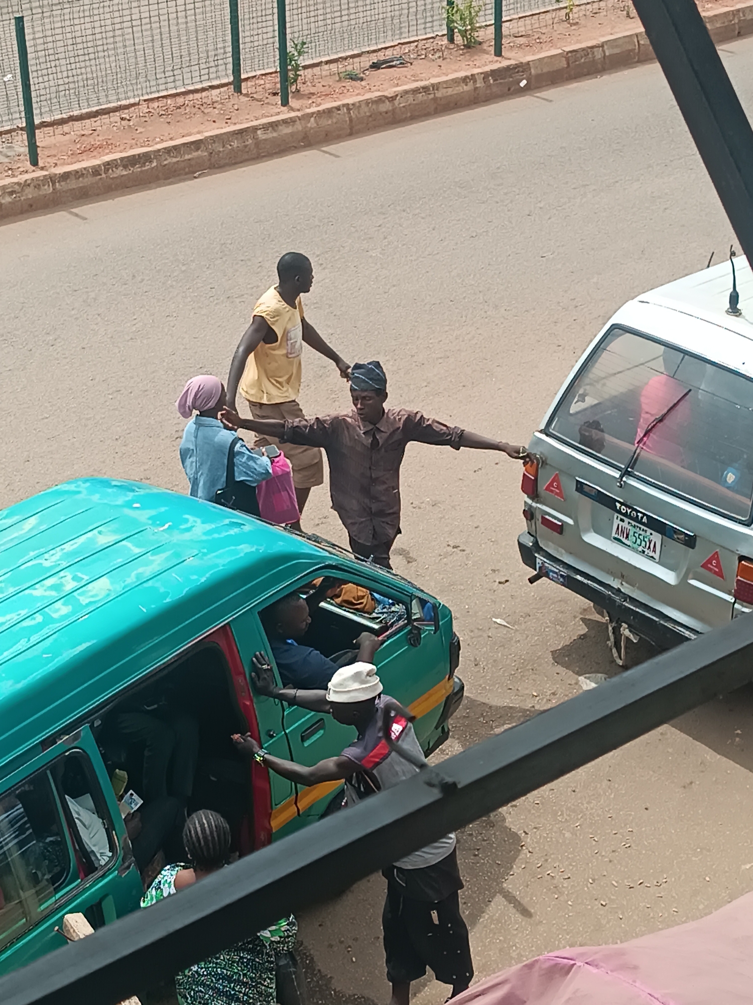 The image shows a street scene in Bukuru, Nigeria. A green bus is parked on the side of the road, with passengers boarding and exiting. A man wearing a white hat and a grey shirt is collecting fares near the bus door. Another silver bus with a license plate reading "ANN 555XA" is parked nearby. Three people stand between the two vehicles. The overall environment is urban, with a fence visible in the background.