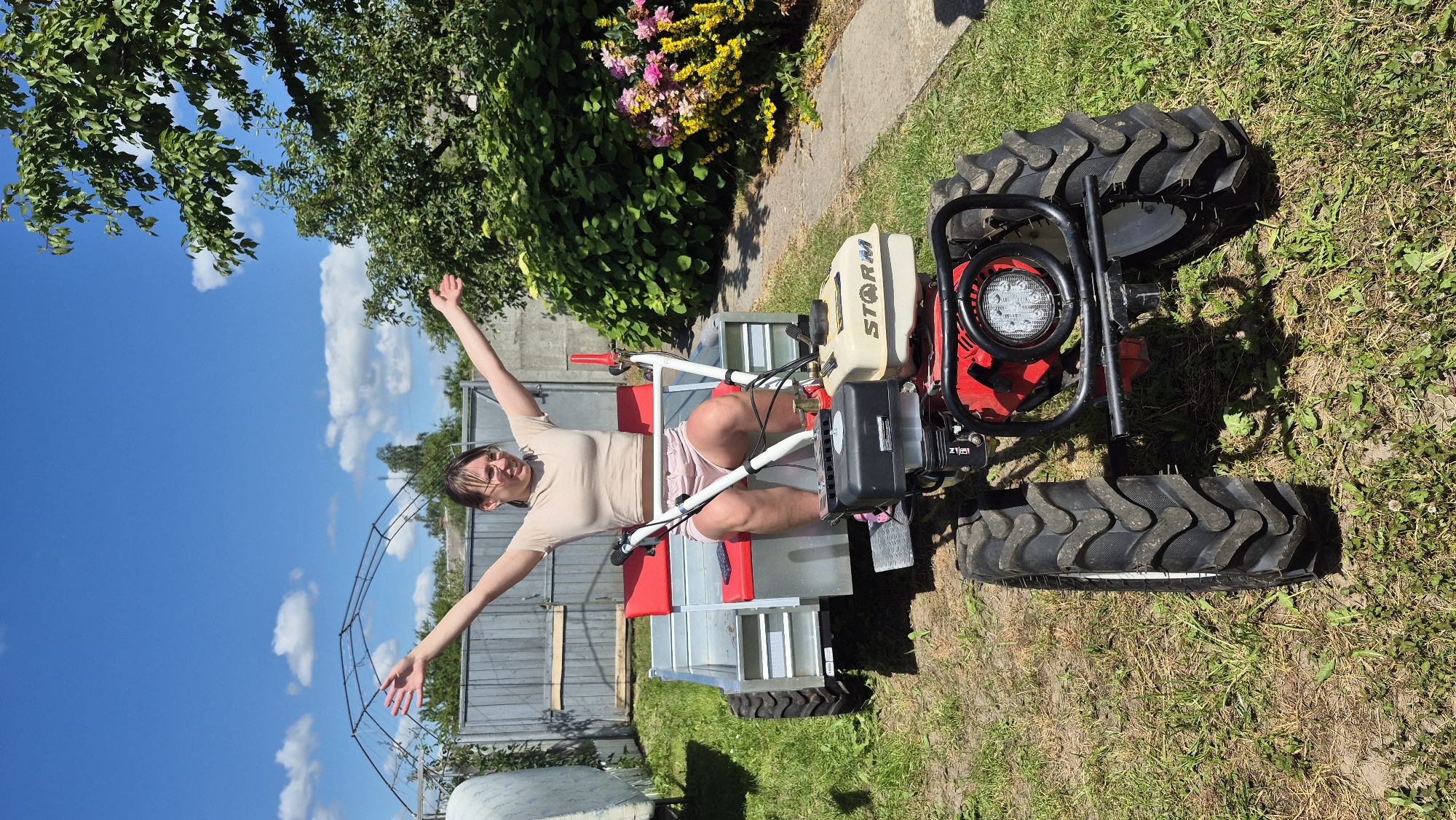 A young woman with dark hair pulled back and glasses is sitting on a small, red and black motor-cultivator with a trailer attached. She is wearing a beige crop top and pink shorts and has her arms raised in a celebratory gesture. The motor-cultivator is parked on a grassy area in front of a chain-link fence and some greenery. In the background, there are trees, a greenhouse-like structure, and a small flowering bush. The sky is blue with some scattered clouds. The location appears to be a rural area, possibly a backyard or garden in Цярэніцкі сельскі Савет, Belarus.