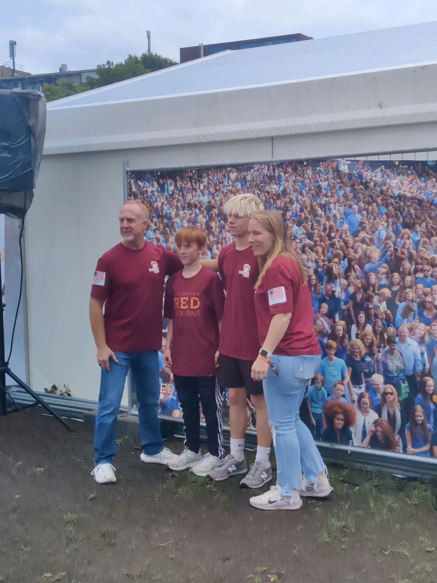 A man, a boy, and a woman are posing for a photo in front of a large mural of a crowd of people. The man is wearing a maroon t-shirt with an American flag patch and blue jeans. The boy is wearing a maroon t-shirt with the words "I'm having a RED hair day!" printed on it. The woman is wearing a maroon t-shirt with an American flag patch and light blue jeans. The mural behind them appears to depict a large gathering of people, mostly wearing blue and red. The ground in front of them is wet and appears to be muddy. In the background, there is a white tent or canopy structure. There are also some trees and buildings visible above the tent. The overall scene suggests an outdoor event. The image does not provide any explicit information to determine the location in the city of Tilburg, Netherlands.