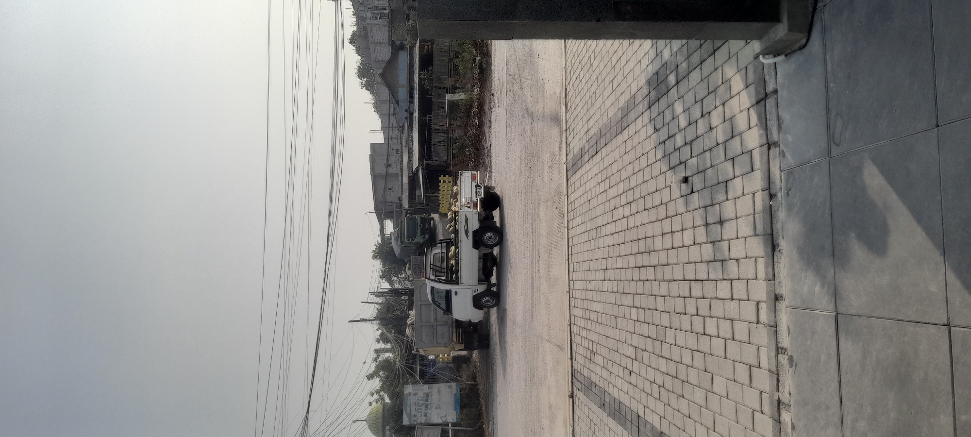A wide shot captures a scene in Kragilan, Indonesia, with a pale sky overhead. In the middle ground, a white pickup truck is parked, its bed filled with round, light-colored objects, likely produce such as melons or coconuts. Behind the pickup truck is a larger, beige truck with a similar load in its open bed. Further back, a green truck is visible, and beyond that, various low-rise buildings and structures. A distinctive green-domed building, possibly a mosque, is seen to the left of the frame, with a tall pole and numerous wires strung between structures. The foreground is dominated by a paved area with a pattern of interlocking square stones. To the right of the frame, a dark, rectangular pillar stands, casting a shadow across the paved area. A discarded plastic cup is visible on the stone paving in the middle ground. The overall environment suggests a commercial or logistical area.
