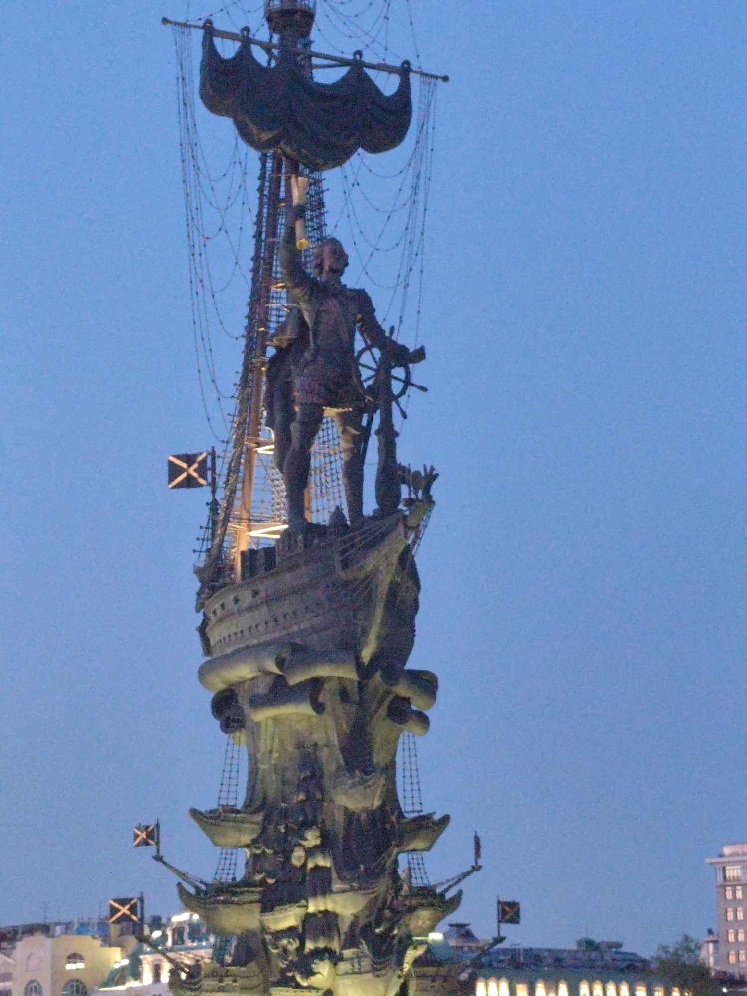 The image depicts a tall statue of a man on a ship, likely a historical figure associated with seafaring. The statue is placed atop a large, ornate column resembling a ship's mast, with multiple levels of ship-like structures extending downwards. Flags bearing an "X" symbol are visible at different points on the structure. The statue and column are bronze or dark metal in color. The background is a clear blue sky, suggesting twilight or dusk. In the lower portion of the image, buildings with lit windows can be seen, indicating that it is an urban environment. The location is the Peter the Great Statue in Moscow, Russia.