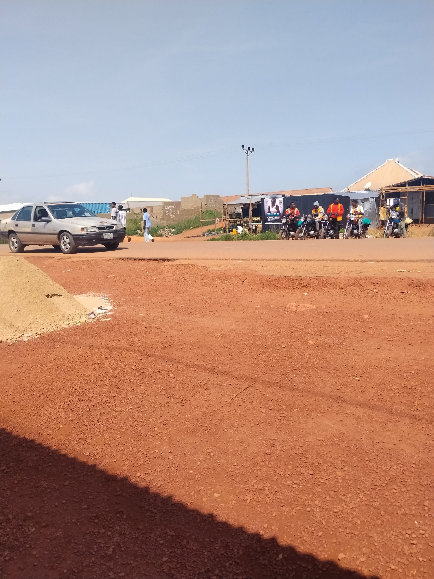 The image captures a street scene in Durumi, Nigeria, under a clear blue sky. A silver sedan is parked on a dirt road, its front tires turned slightly to the left. A small pile of light-colored dirt is visible to the left of the car. In the background, several individuals are standing or walking near a cluster of low-rise buildings with corrugated metal roofs. A large poster featuring individuals and text is displayed on the facade of one of the buildings. Further down the street, a line of motorcycles with riders is parked. The overall environment is dusty and appears to be a mixed residential and commercial area.