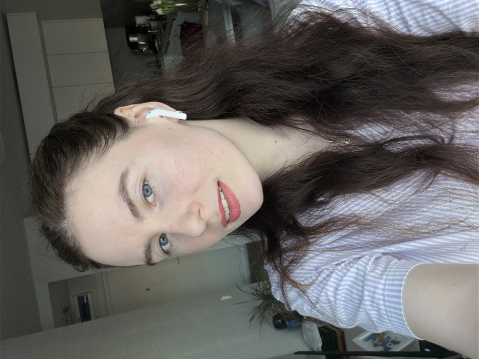 The scene is located indoors in the city of Ковалёво, Russia. A fair-skinned female, appearing to be in her mid-20s to early 30s, is positioned in the foreground, facing directly towards the camera. She has long, wavy dark brown hair, parted on the side and partially pulled back, and light blue eyes. Her lips are colored with light pink lipstick, and she exhibits a slight smile. A white wireless earbud is inserted into her right ear. She wears a light purple and white striped short-sleeved collared shirt. A delicate necklace with a small, clear or light-colored pendant is visible around her neck.
The background indicates a residential interior. To the subject's left, a light-colored wall is visible, along with a dark, upright object, possibly a door frame, and a partial view of a green potted plant. To the subject's right, a modern kitchen area is discernible, featuring white upper cabinets and a dark countertop. A vase containing several white flowers, consistent with tulips, is placed on the countertop, alongside other indistinct objects. The lighting in the scene is bright and consistent.