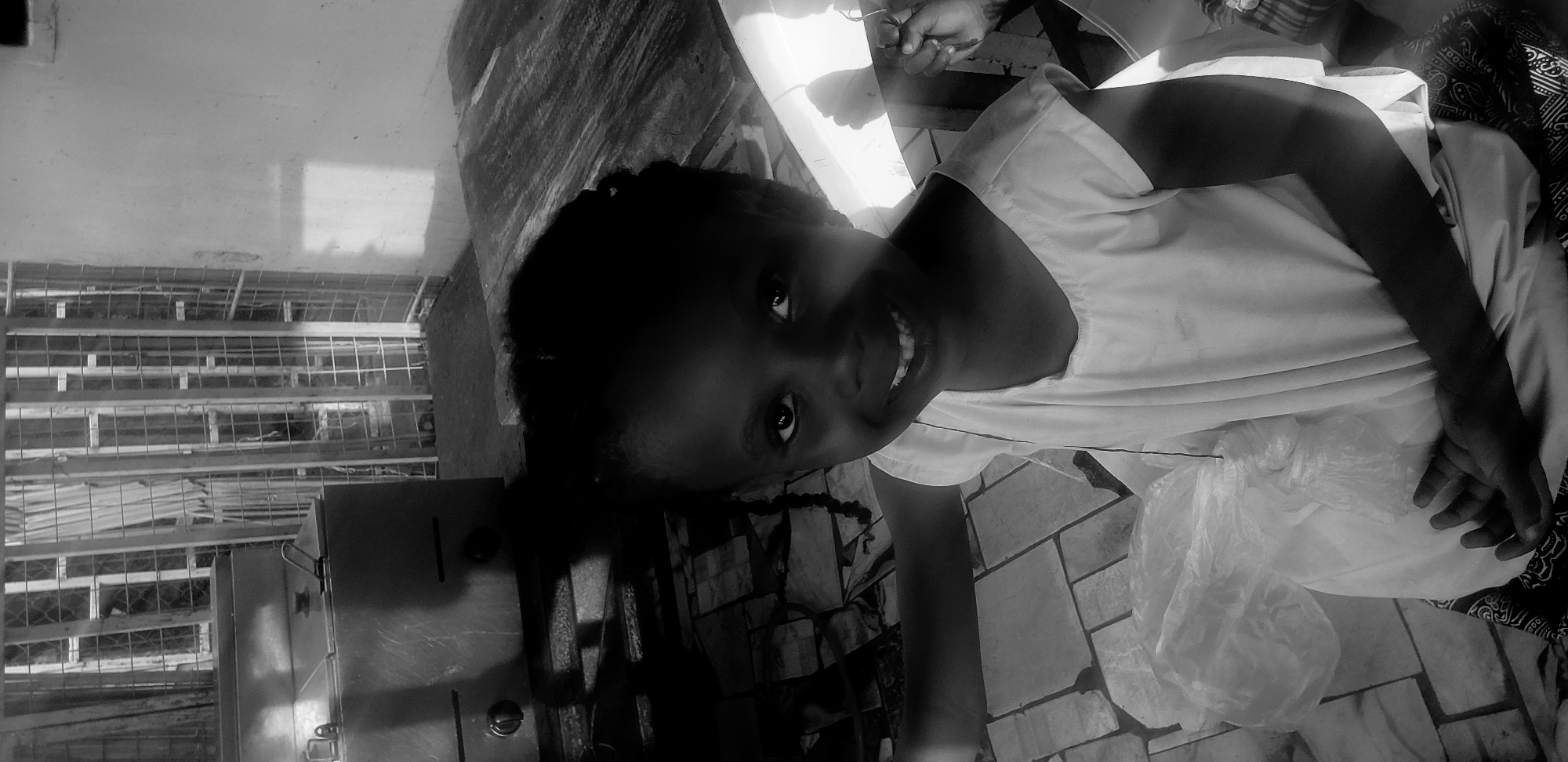 A black and white, eye-level medium shot captures a young Black girl with braided hair, smiling directly at the camera. She is seated and wearing a simple, sleeveless white dress that drapes over her lap. To her left, a crumpled white plastic bag rests on the floor. The background is busy, with a stainless steel appliance, possibly a deep fryer, to the left, and a wooden table to the right. Behind these, a latticed structure, possibly a window or gate, is visible. The floor is tiled with a mosaic pattern. The setting appears to be an outdoor or semi-enclosed area, with sunlight casting shadows on a plastered wall. The overall impression is of a candid moment, with the girl's bright smile being the focal point. This scene is located in Narayi, Nigeria.