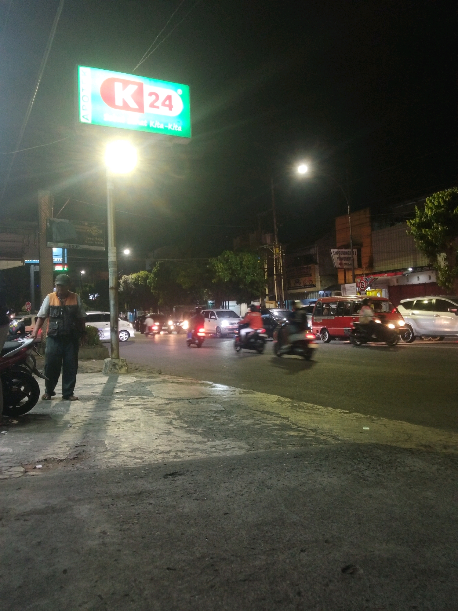 This is a nighttime, eye-level, medium shot of a street scene in Purwokerto, Indonesia. A man wearing a vest over a light-colored shirt and dark pants stands on a sidewalk near a streetlamp. The street is busy with traffic, including motorcycles, cars, and a red van. A prominent illuminated sign for "K 24" with the text "APOTEK" and "Kita-Kita" is visible in the upper left quadrant of the image. The street is paved and appears slightly worn, with some patches and cracks. Trees and buildings line the street in the background. The overall lighting is dim, with the streetlamp and vehicle headlights providing most of the illumination.
