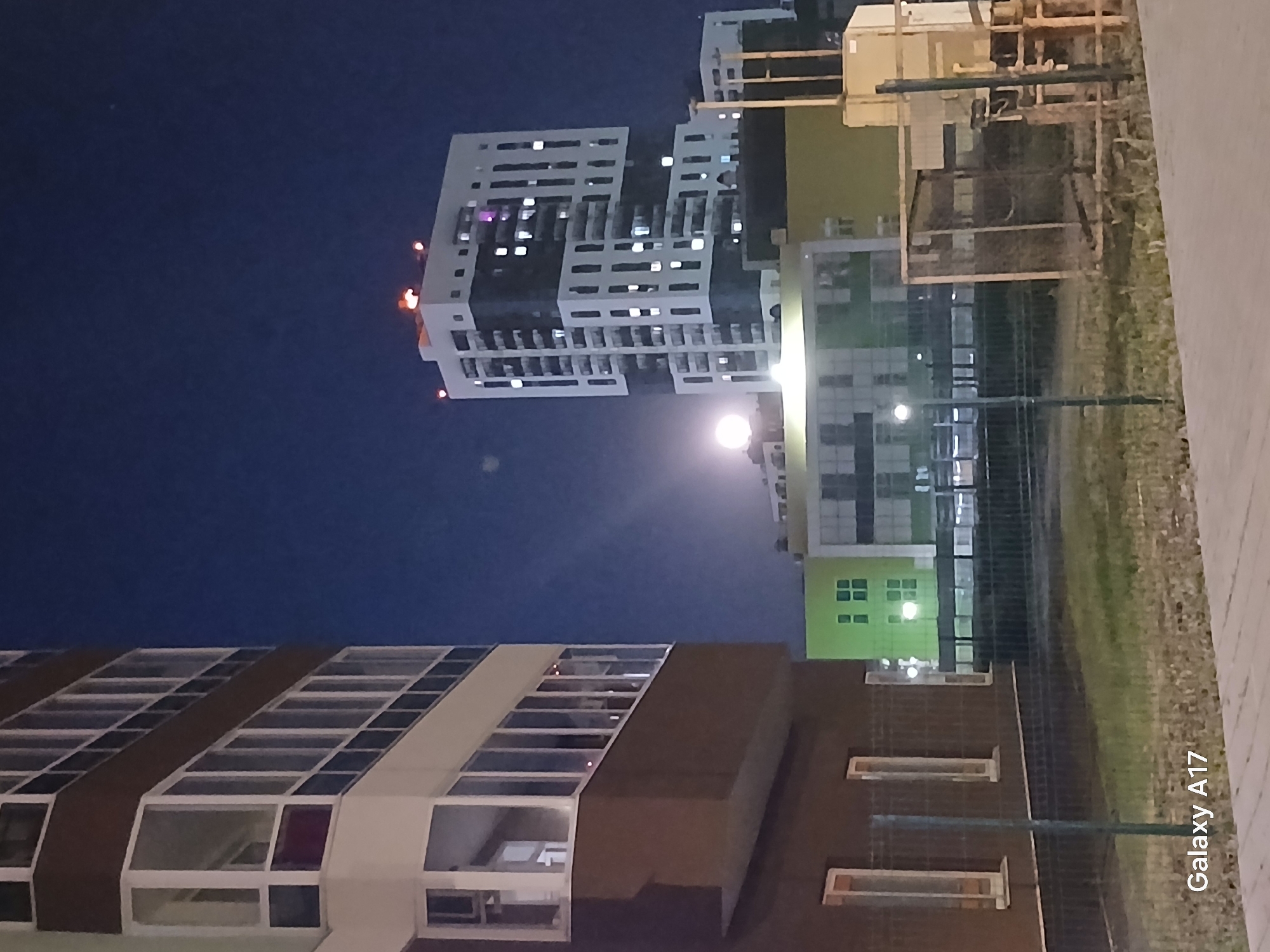 The image shows a nighttime view of buildings in Yekaterinburg, Russia. A tall, modern building with a white facade and illuminated windows stands prominently in the upper part of the frame. A lower, brown building with a pitched roof and skylights is visible in the lower part. Streetlights illuminate the area between the buildings. The sky is dark blue. There are some wooden constructions behind a fence.