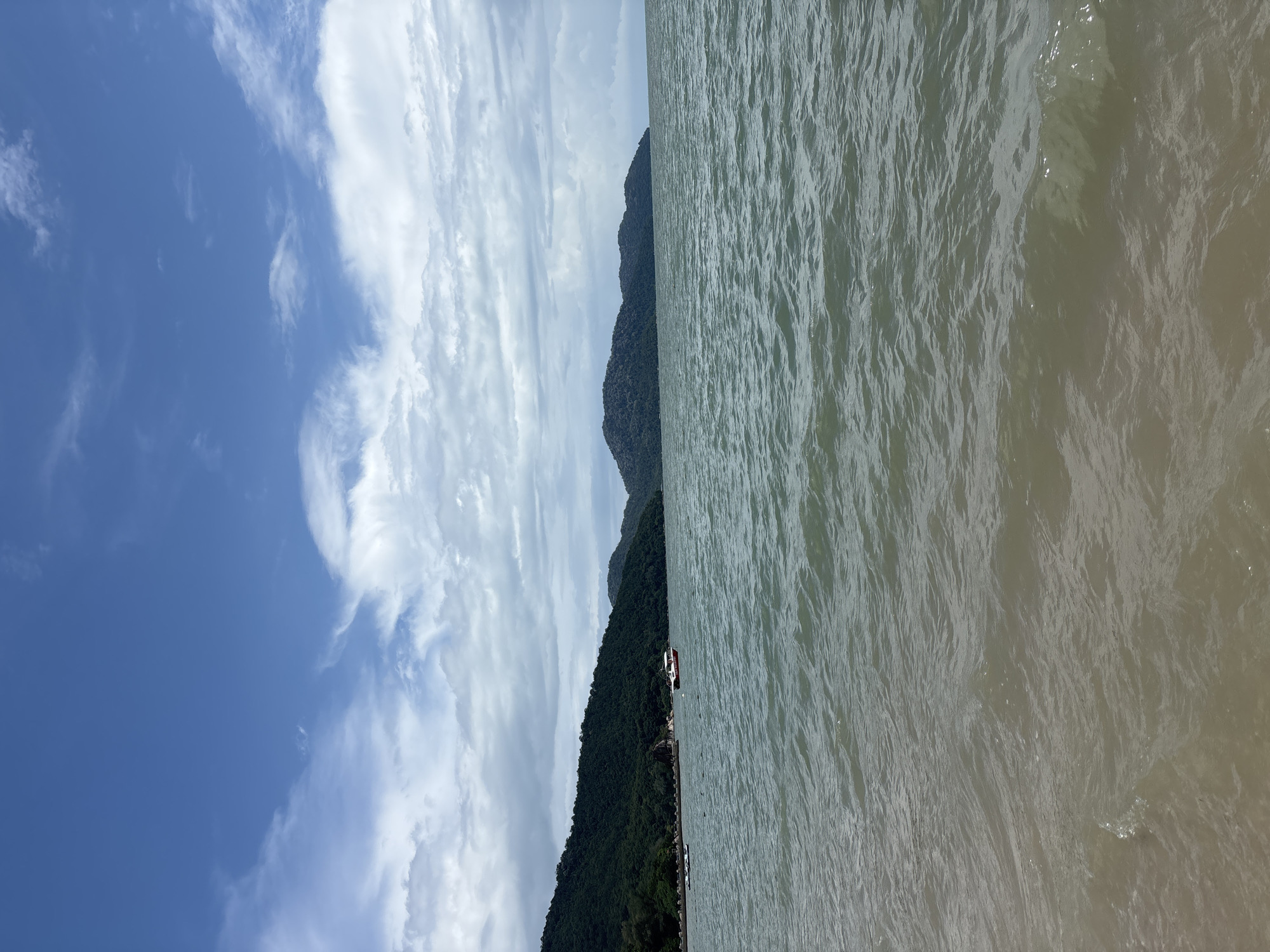 The image depicts a serene view of the sea with a coastline covered in lush green vegetation in the background. The water is calm with gentle ripples, reflecting the overcast sky. Two boats are visible in the water, one a red and white motorboat, and another smaller, white boat closer to the shore. The sky is a mix of blue and white clouds, with a few wisps of cirrus clouds adding texture. The overall scene evokes a sense of tranquility and natural beauty. The image was taken in the city of George Town, Malaysia, likely from a beach or a coastal area.