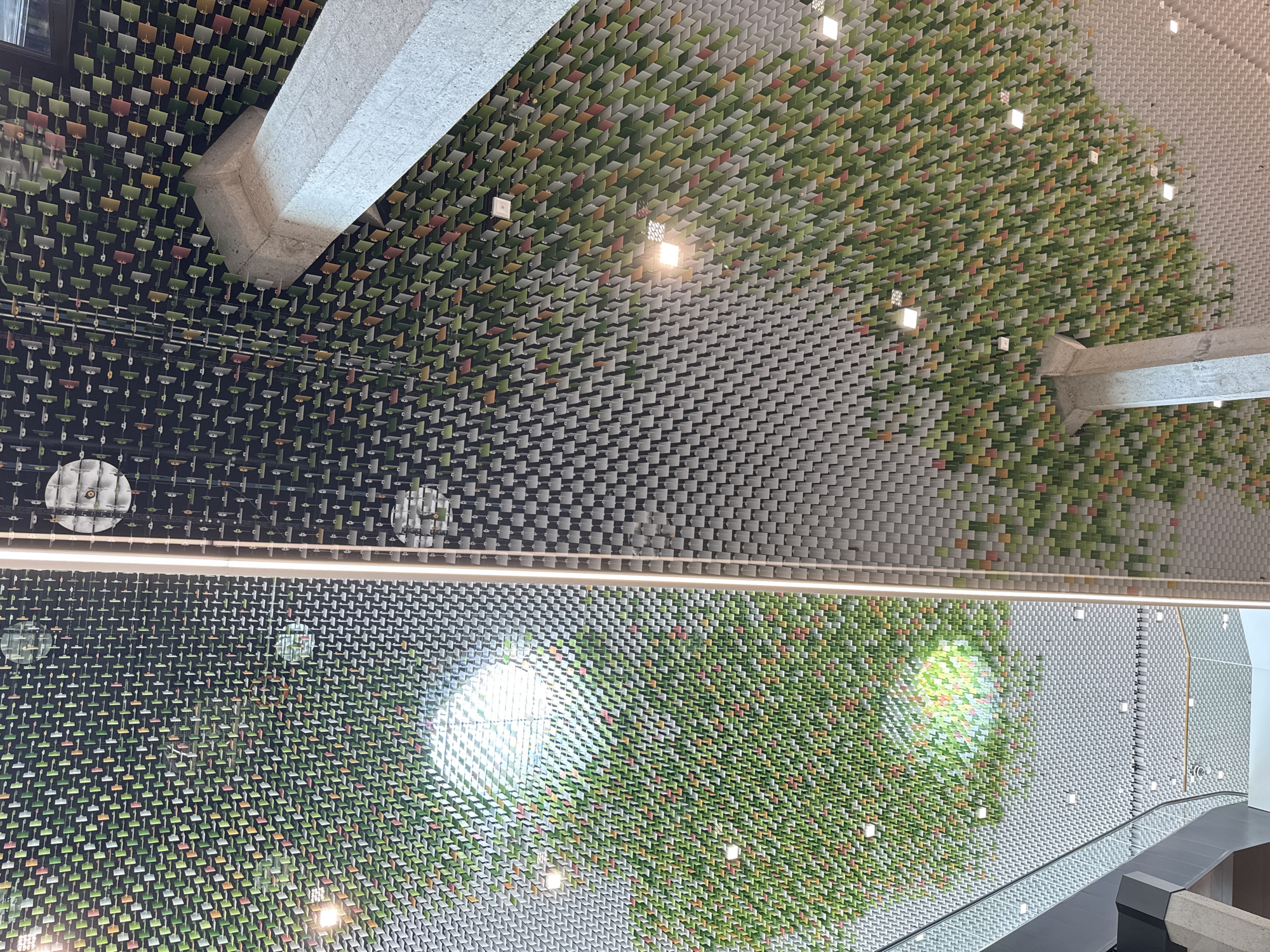 The image shows an interior view of a ceiling in the city of Morbio Inferiore, Switzerland. The ceiling is a complex structure composed of numerous small, angled panels. These panels are arranged in a repetitive pattern, creating a textured surface. Some sections of the ceiling feature panels in various shades of green, yellow, and red, suggesting a decorative or artistic element. Other sections are predominantly grey or black.
Several light fixtures are visible, some recessed and square, others circular and protruding. A prominent concrete pillar, possibly structural, is visible in the foreground and mid-ground, extending upwards into the ceiling. The overall impression is of a modern architectural design, potentially a public space or a commercial building, characterized by its intricate ceiling treatment and the interplay of light and shadow. Reflections are visible on some of the panels, indicating a polished or reflective surface. A glass railing is also visible in the lower left portion of the image, suggesting a mezzanine or elevated walkway.