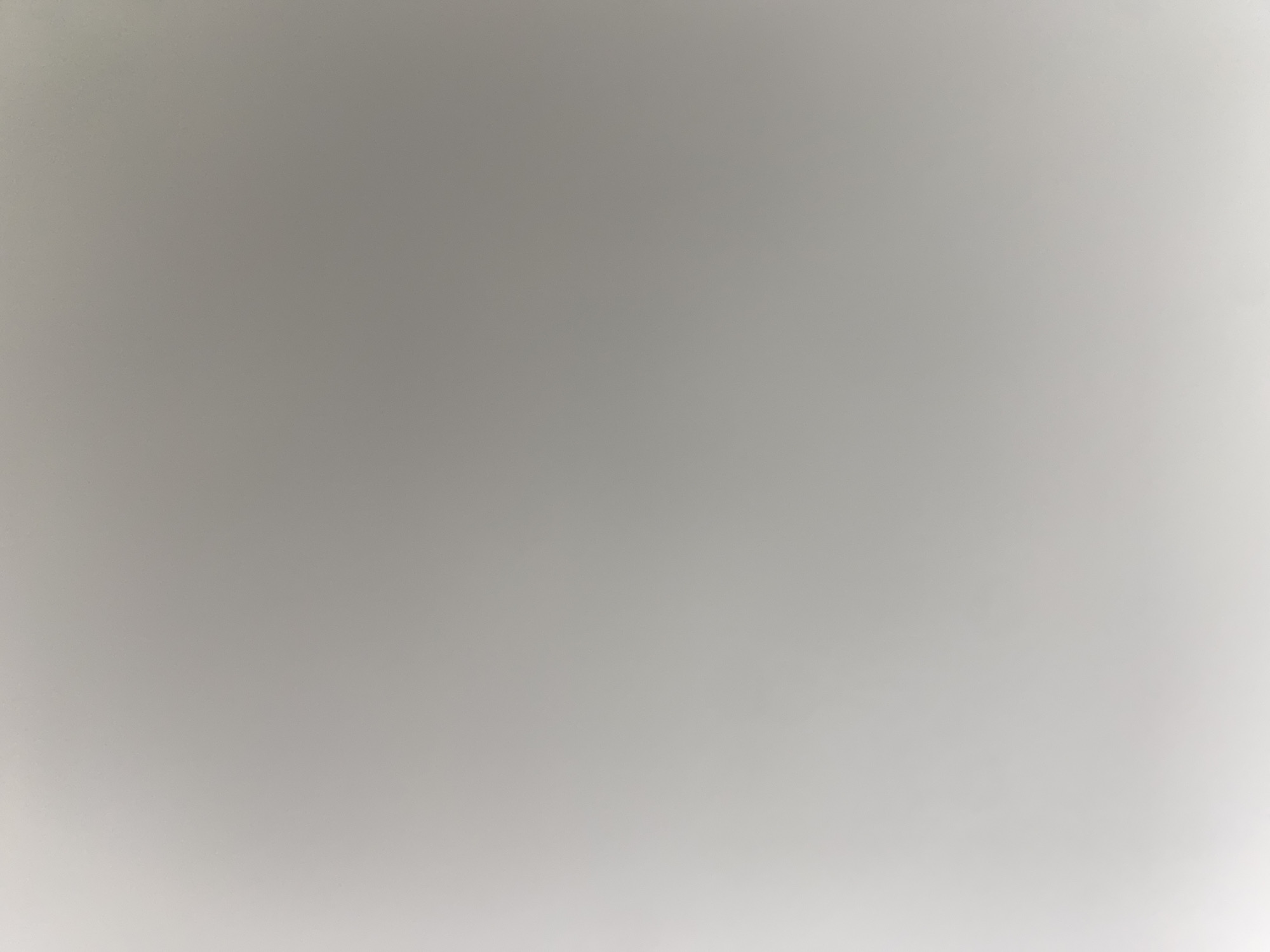 The image is a close-up, out-of-focus shot of a plain, light gray surface. There are no discernible objects, people, or environmental details. The lighting appears to be uniform, with a slight gradient from darker at the top to lighter at the bottom. The image lacks any specific context or identifiable features, making it impossible to determine a location within a city or any other setting.