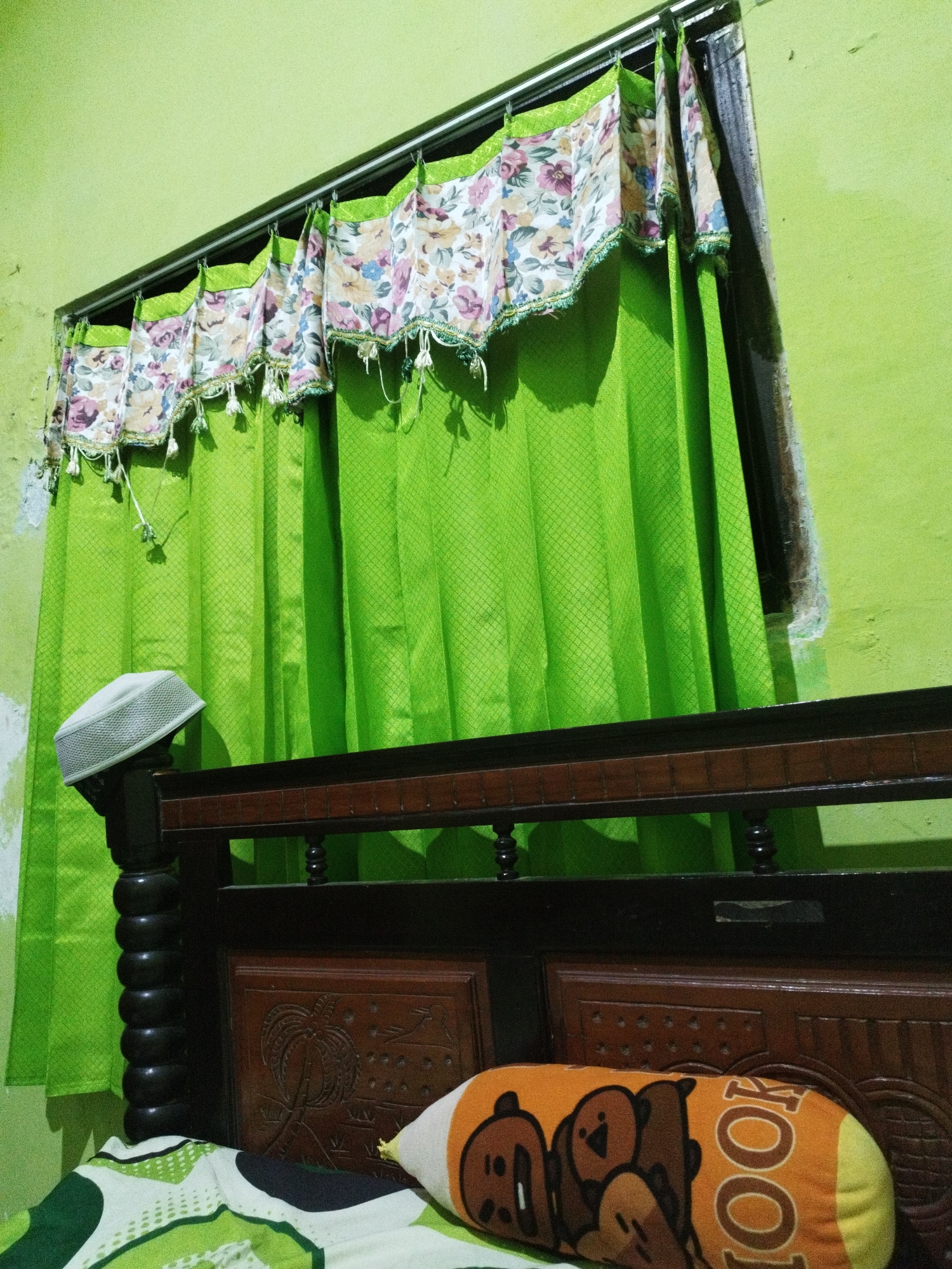 A white knit cap rests on the dark wooden headboard of a bed. Behind the headboard, bright green curtains with a diamond pattern are drawn across a window. A decorative valance with a floral pattern hangs above the curtains. A cylindrical pillow with cartoon bear faces and the word "LOOK" printed on it lies on the bed. The room has a light green wall. The scene is located in Marengan Daya, Indonesia.