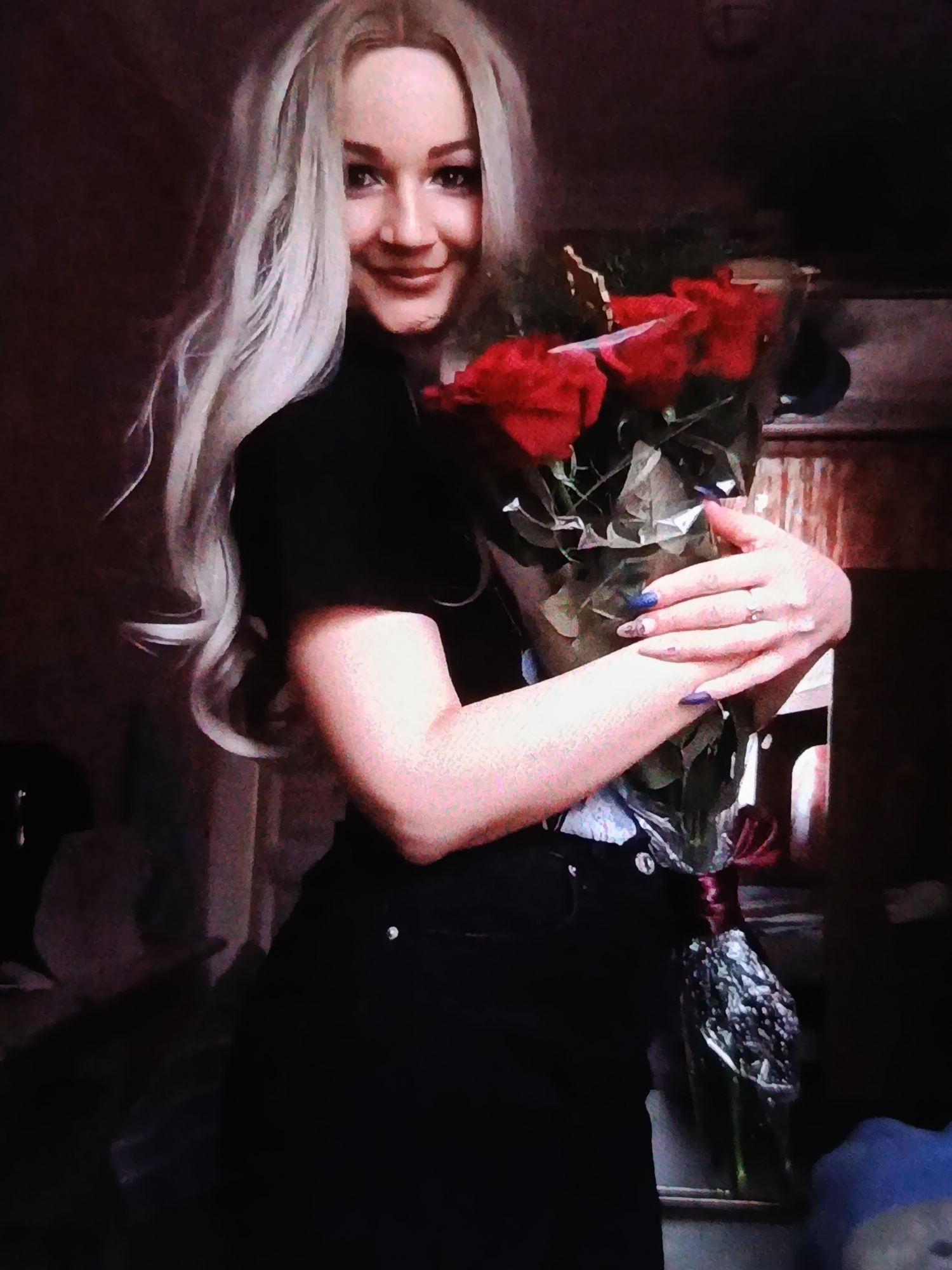 A medium-shot, full-body photograph shows a young woman with long, blonde hair holding a bouquet of red roses. She is wearing a black top and black pants. Her left arm is wrapped around the bouquet, and her right hand rests on top of it. The woman is looking at the camera and smiling. The background is dark and out of focus, with some hints of furniture and decor. The lighting is dim, with a spotlight on the woman and the roses. The photograph appears to have been taken indoors.