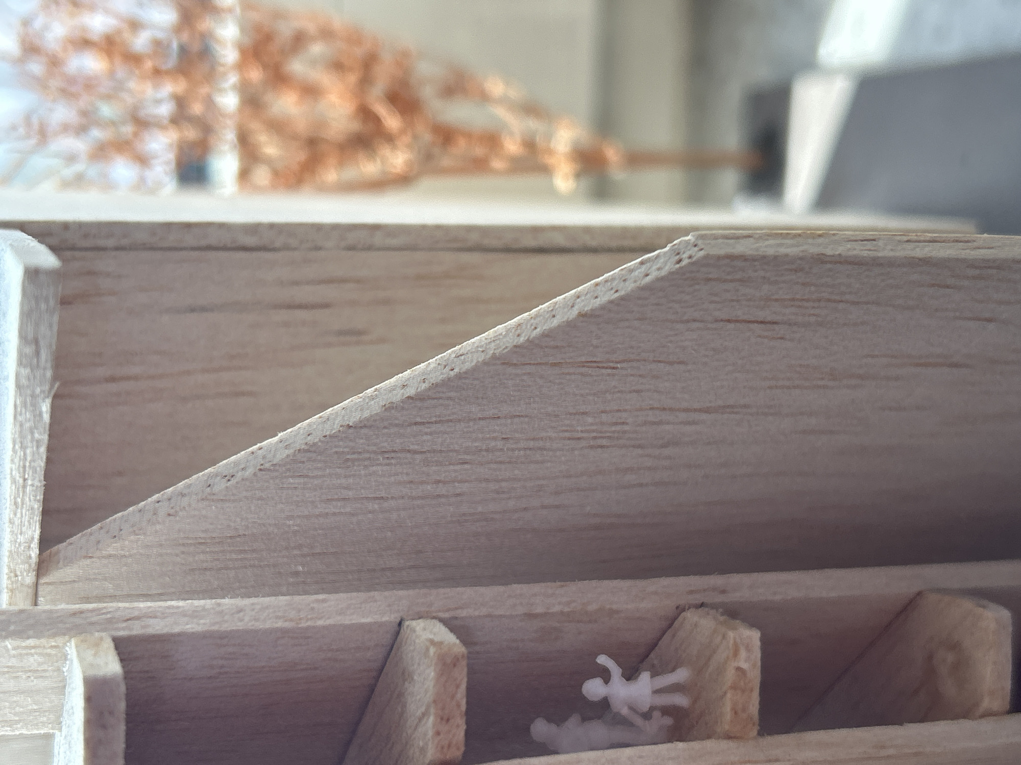 The image shows a close-up of a small, unfinished wooden structure. The structure appears to be divided into compartments. Two small, white figurines are placed in one of the compartments. The wooden pieces show wood grain and rough cut edges. The background is blurred, with hints of dried flowers and an indistinct white structure. The location is not discernible from the image.