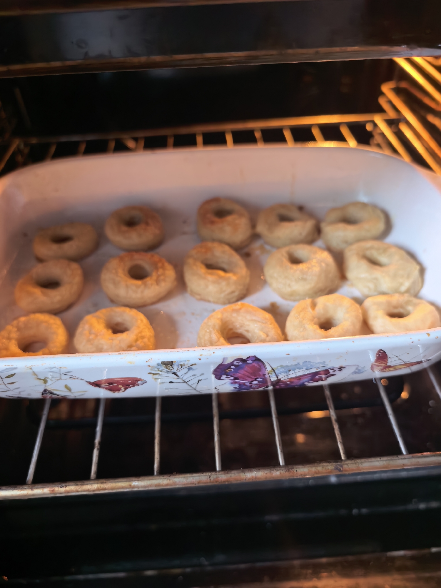 The image captures a close-up view inside a dark oven, where a white rectangular baking dish holds approximately 15-18 small, ring-shaped pastries, likely donuts or small bagels. The pastries, arranged in rows, appear light golden brown, indicating they are in the process of baking or toasting.
The baking dish features a decorative rim with colorful botanical and insect motifs, including a prominent red and purple butterfly on the front edge, along with green and brown leaves and smaller red accents. The dish rests on a metal oven rack. Above the dish, the oven's upper heating element glows with a bright orange light, illuminating the scene and suggesting the oven is actively heating. Other metal oven racks are visible above and below the dish. The overall setting is a kitchen in Sterlitamak, Russia, as suggested by the location context, though no specific geographical cues are visible within the image itself. There is no visible text. The scene conveys a sense of domestic activity, specifically food preparation.